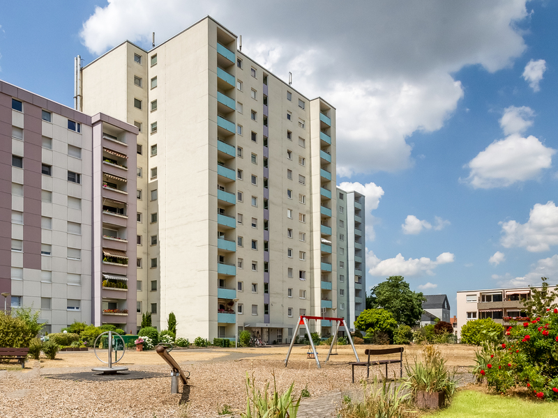 Mehrstöckiges Wohnhaus mit Spielplatz und Sitzbank im Vordergrund.