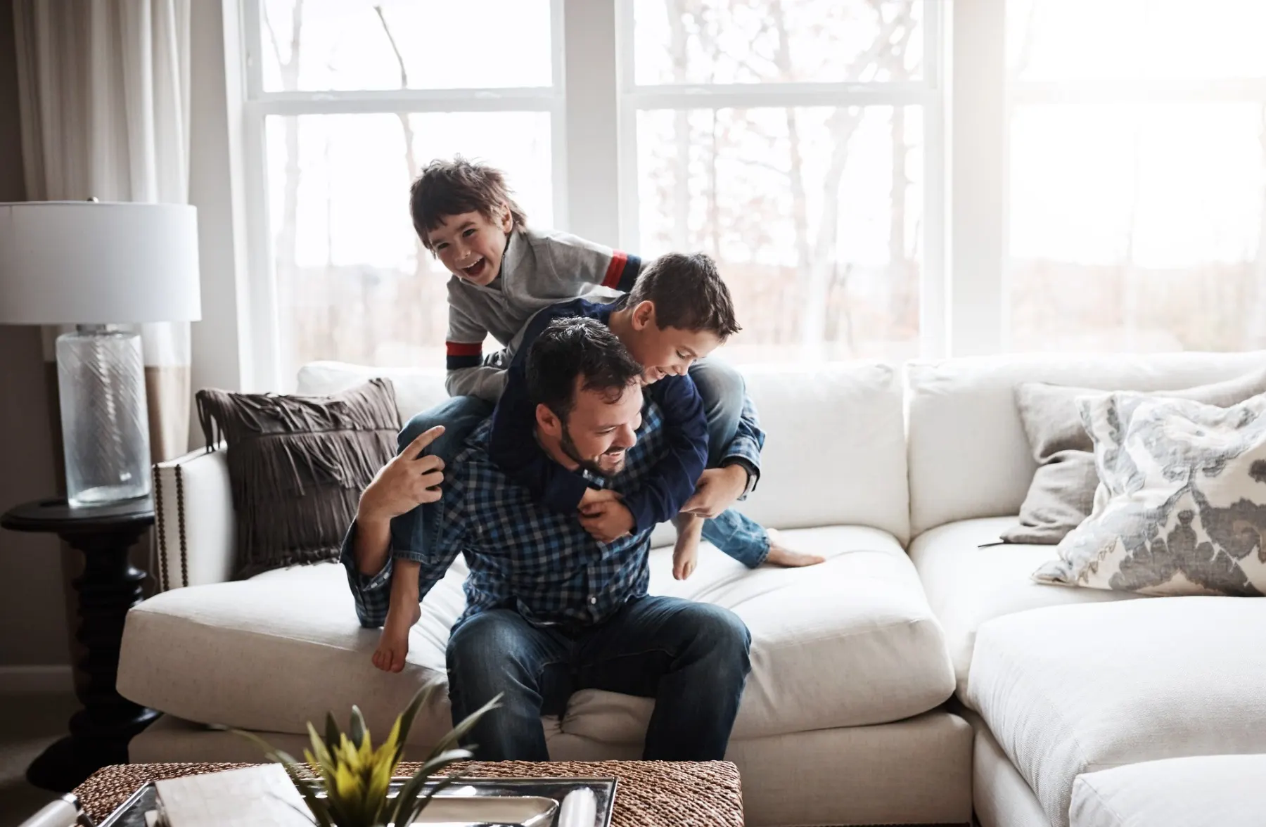Ein Mann spielt mit zwei Kindern auf einem hellen Sofa in einem Wohnzimmer mit großen Fenstern.