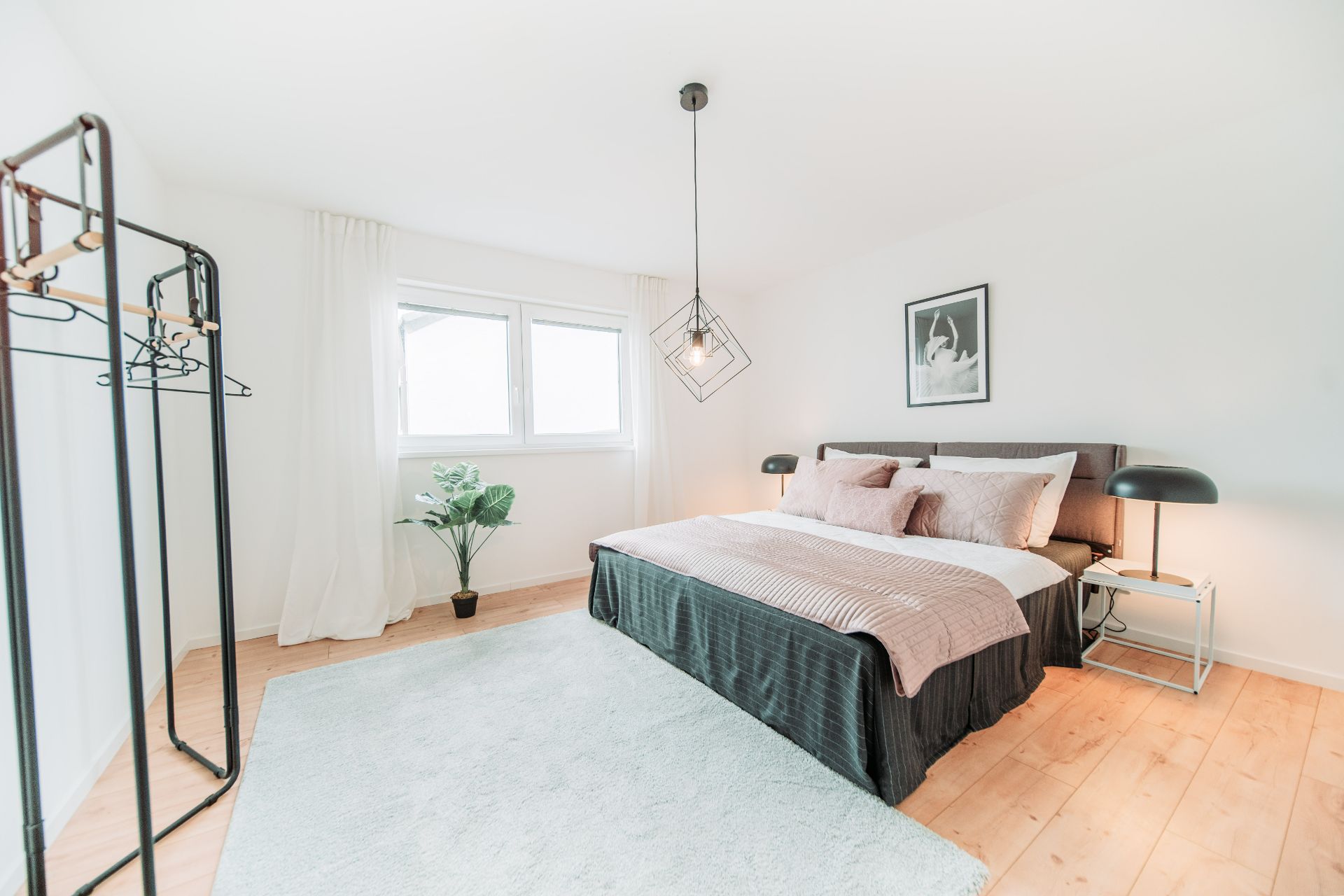 Helles Schlafzimmer mit Doppelbett, Teppich, Kleiderständer, Pflanze und großem Fenster.