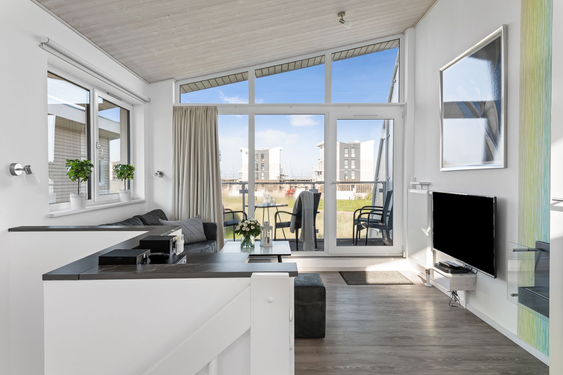 Helles Wohnzimmer mit Sofa, Fernseher, großem Fenster und Zugang zu einem Balkon mit Sitzgelegenheiten.