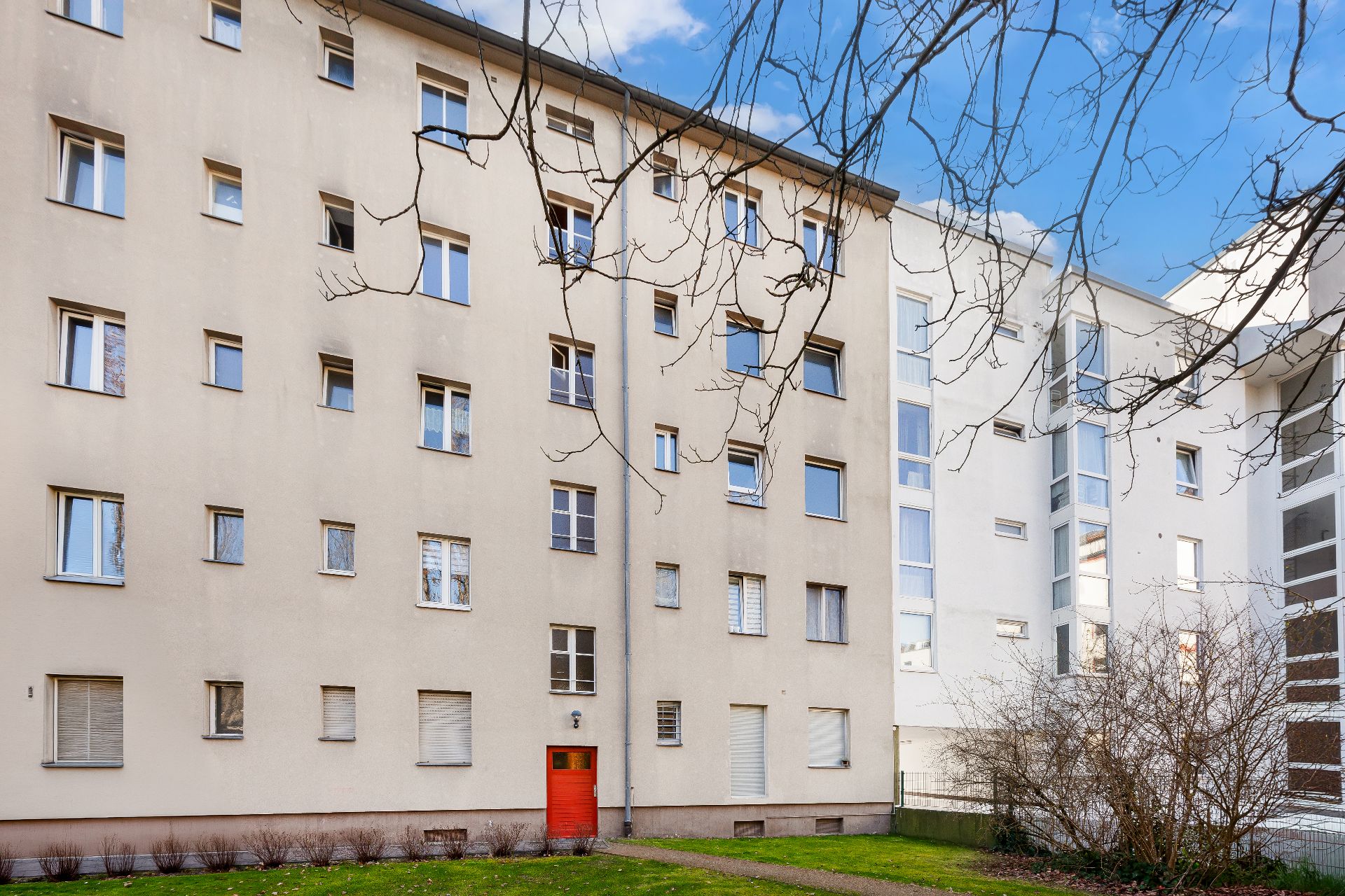 Mehrstöckiges Wohngebäude mit vielen Fenstern und roter Eingangstür, davor eine Rasenfläche.