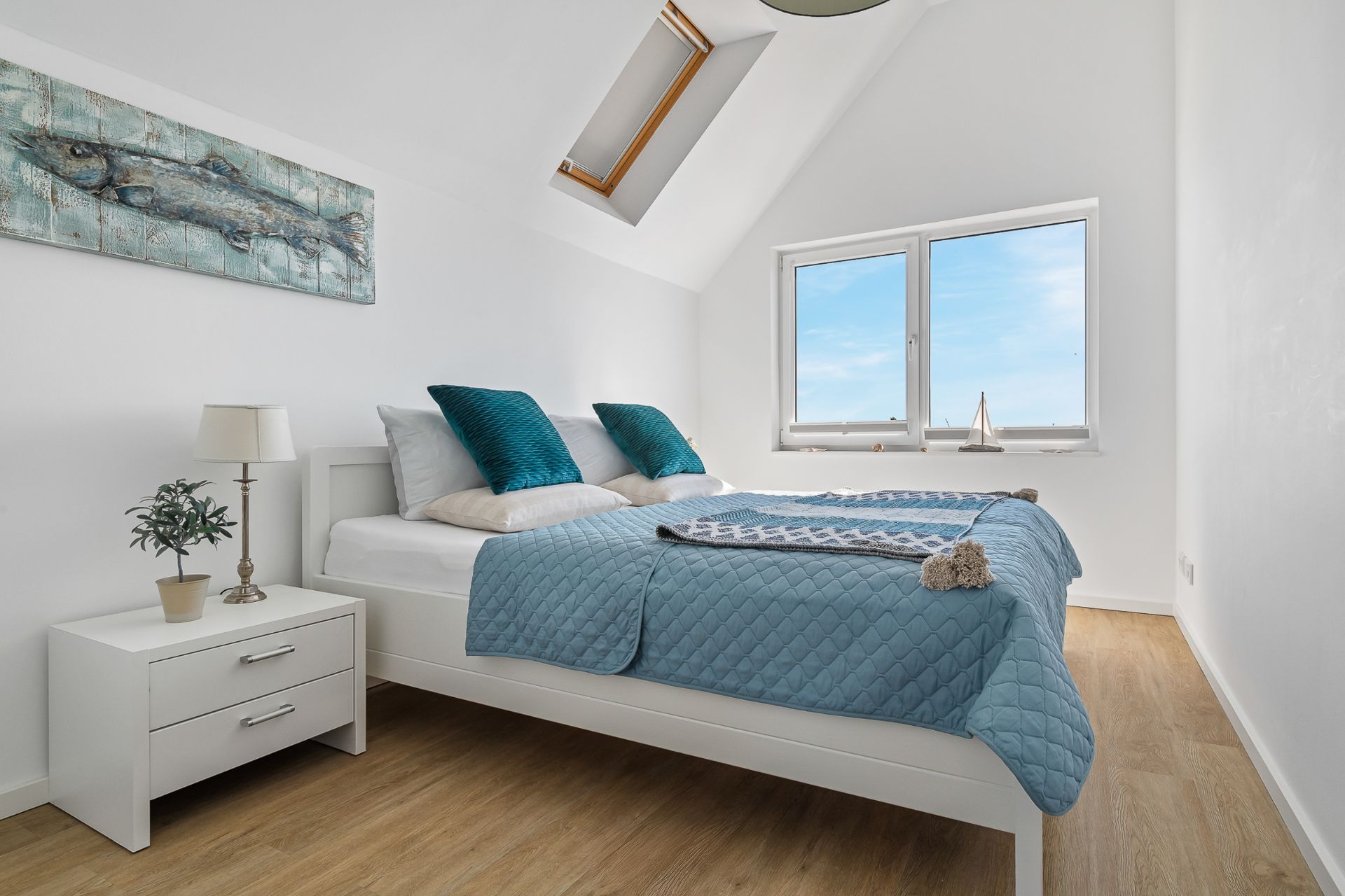 Helles Schlafzimmer mit Doppelbett, blauer Decke, Nachttisch und großem Fenster.