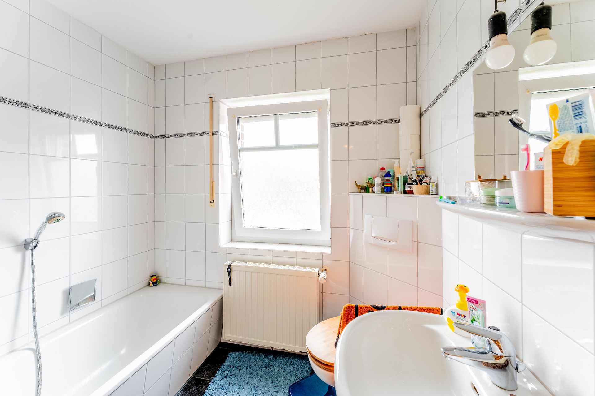 Helles Badezimmer mit Badewanne, Fenster, Waschbecken und Regal voller Pflegeprodukte.
