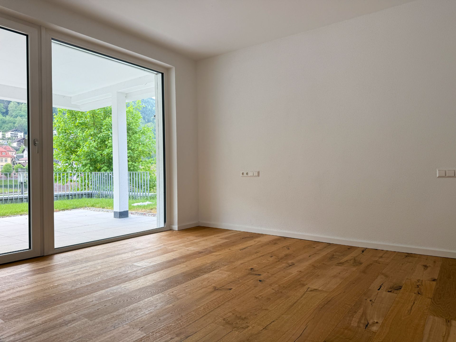 Leeres Zimmer mit Holzfußboden und großer Glastür zu einer Terrasse mit Blick ins Grüne.