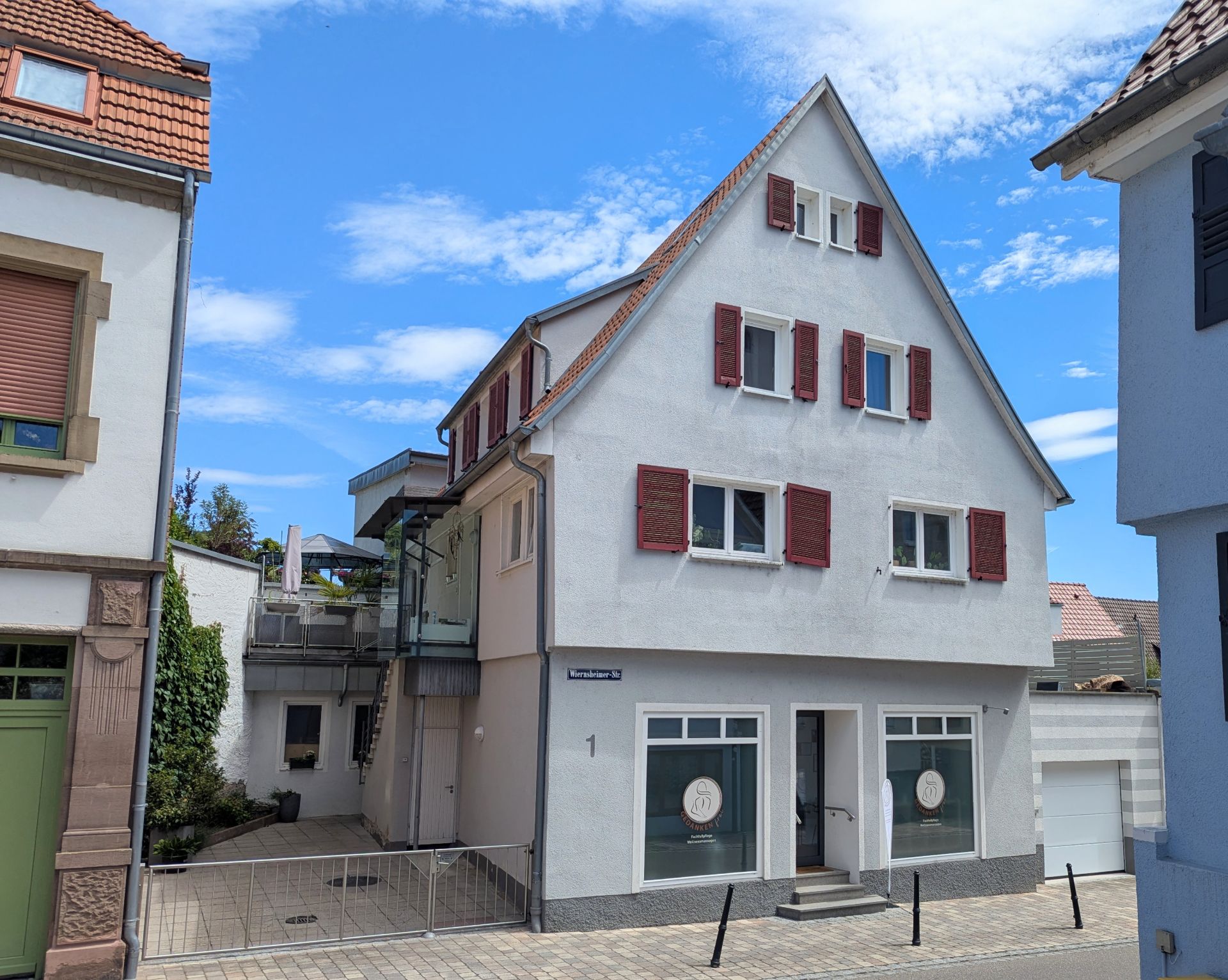 Zweistöckiges Wohnhaus mit rotem Dach, weißen Wänden und roten Fensterläden an einer Straße.