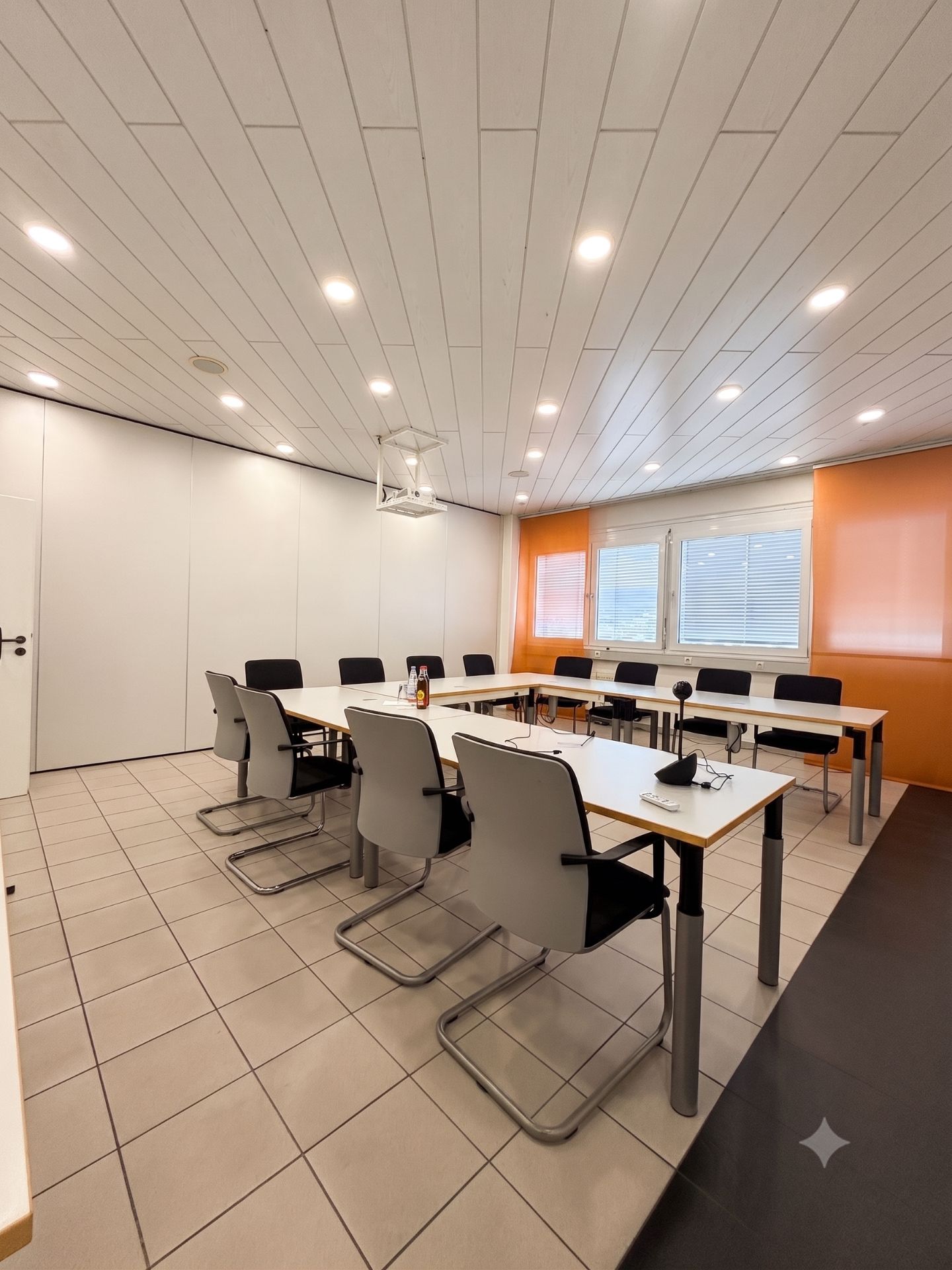 Heller Raum mit mehreren Tischen und Bürostühlen, Fenster mit Jalousien, orange Wand.