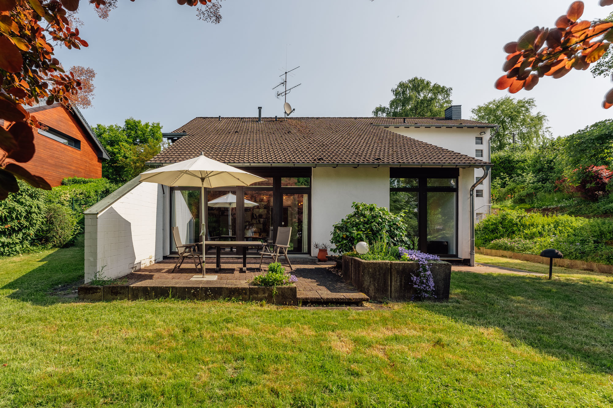Haus mit Terrasse, Tisch, Stühlen und Sonnenschirm, umgeben von Garten und Pflanzen.