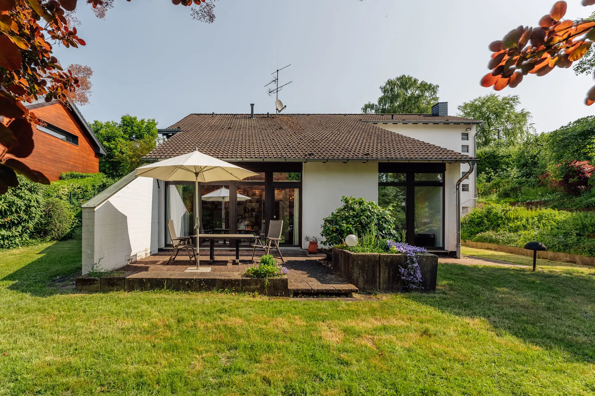Haus mit Terrasse, Tisch, Stühlen und Sonnenschirm, umgeben von Garten und Pflanzen.