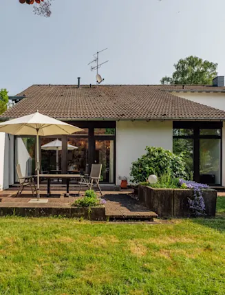 Haus mit Terrasse, Tisch, Stühlen und Sonnenschirm, umgeben von Garten und Pflanzen.