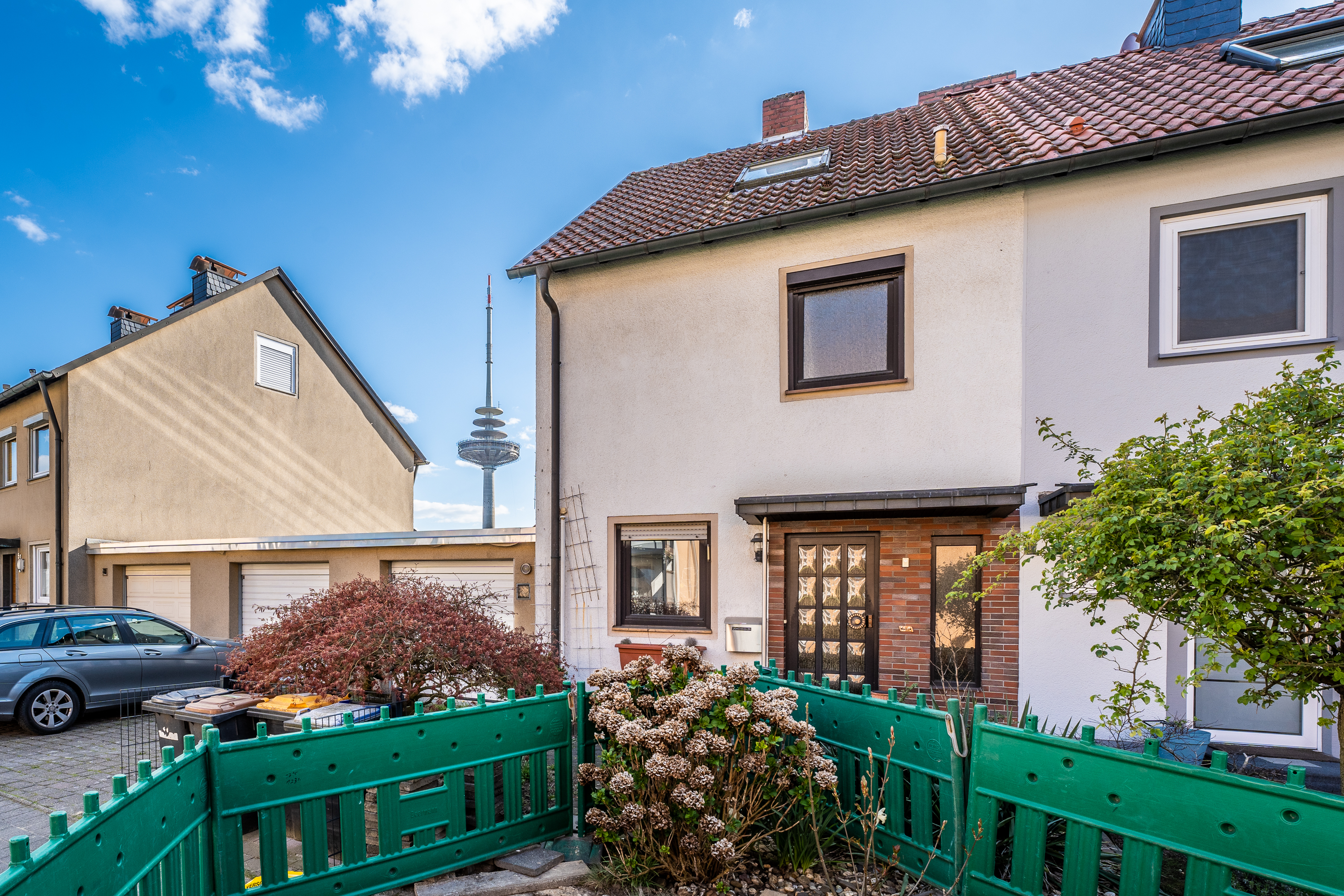 Zweistöckiges Wohnhaus mit grünem Zaun, Vorgarten und Blick auf einen Fernsehturm.