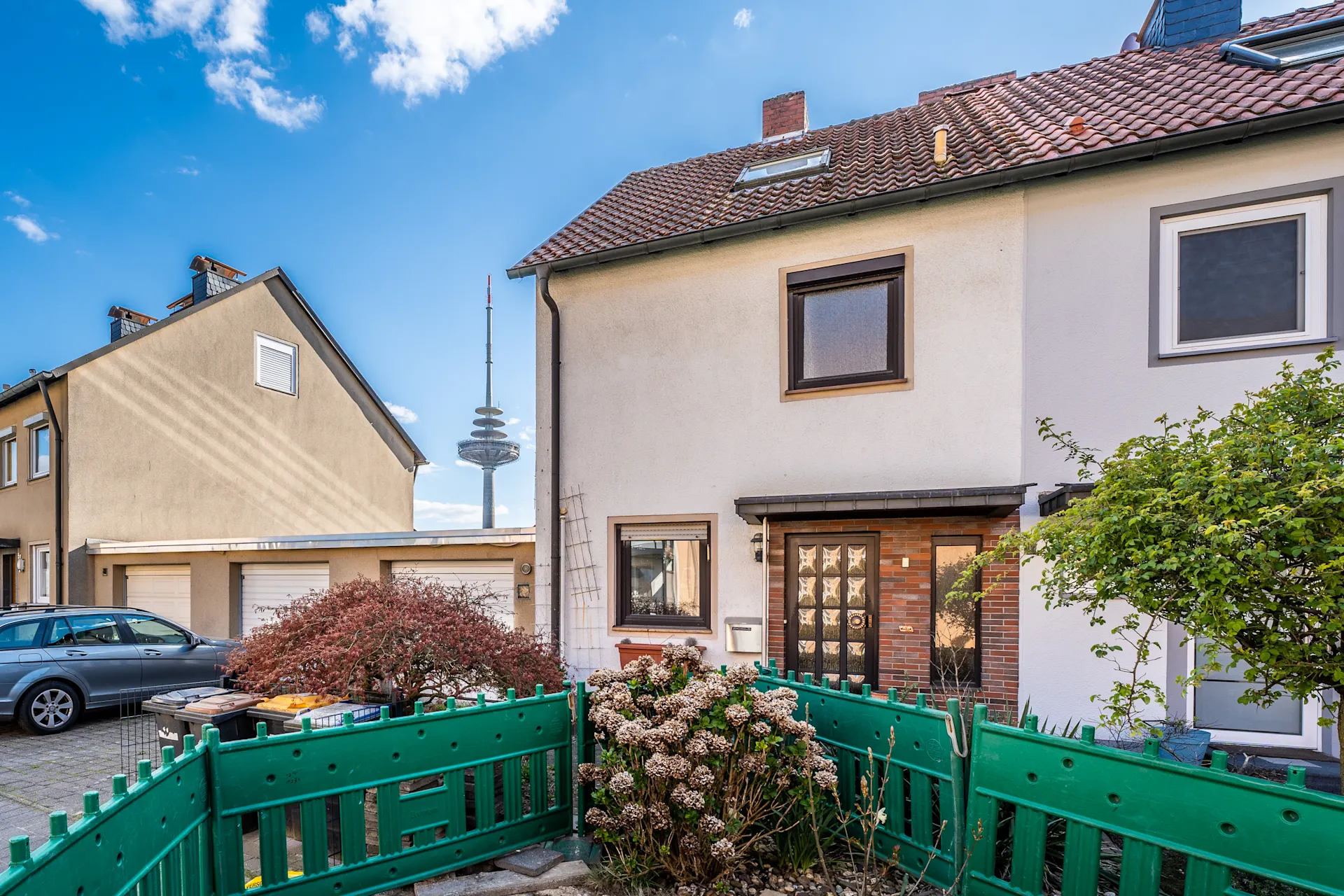 Zweistöckiges Wohnhaus mit grünem Zaun, Vorgarten und Blick auf einen Fernsehturm.