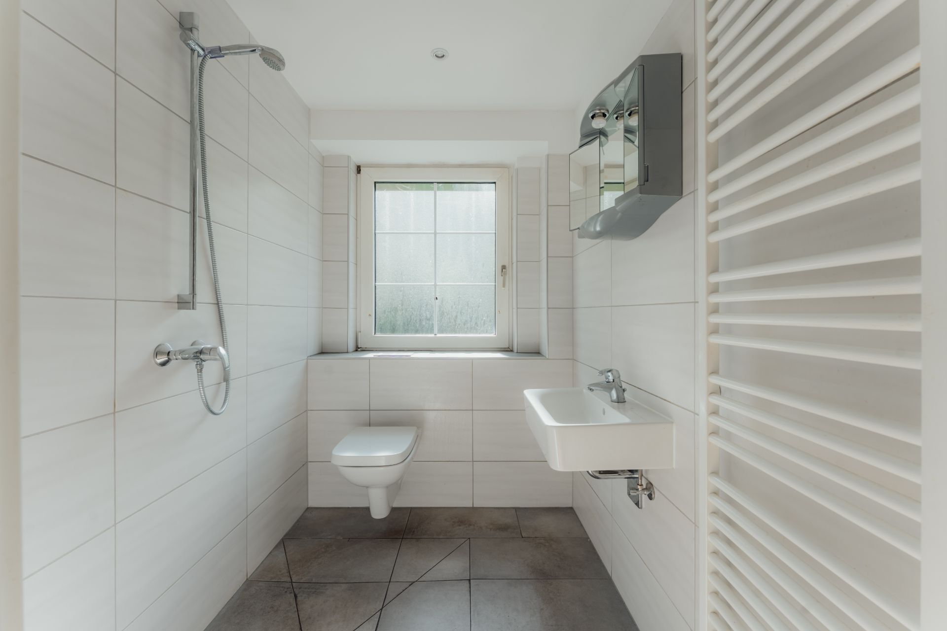 Modernes Badezimmer mit Dusche, Wandtoilette, Waschbecken, Fenster und Heizkörper.