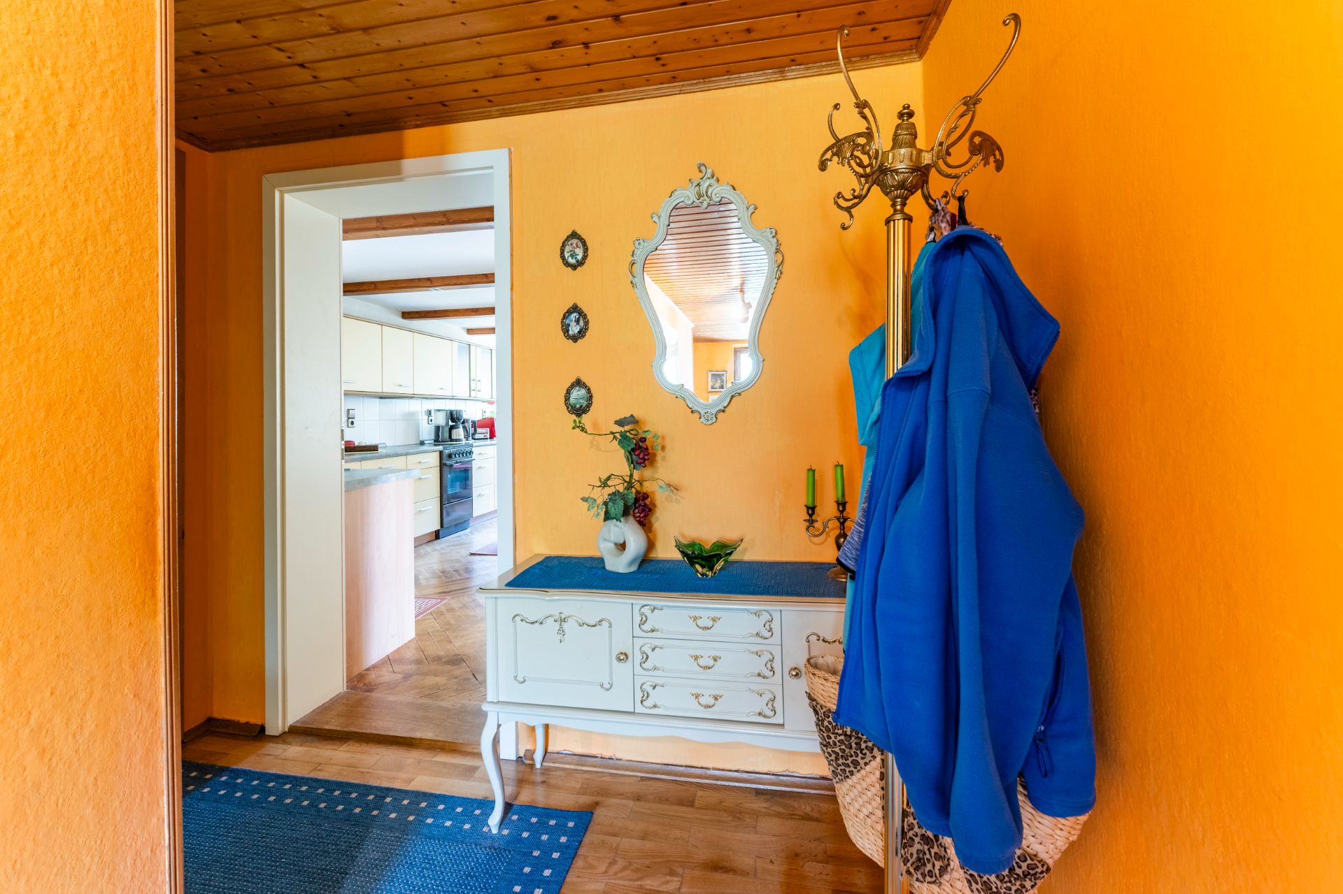 Flur mit orangener Wand, Spiegel, Kommode, Garderobe mit blauer Jacke und Blick in die Küche.