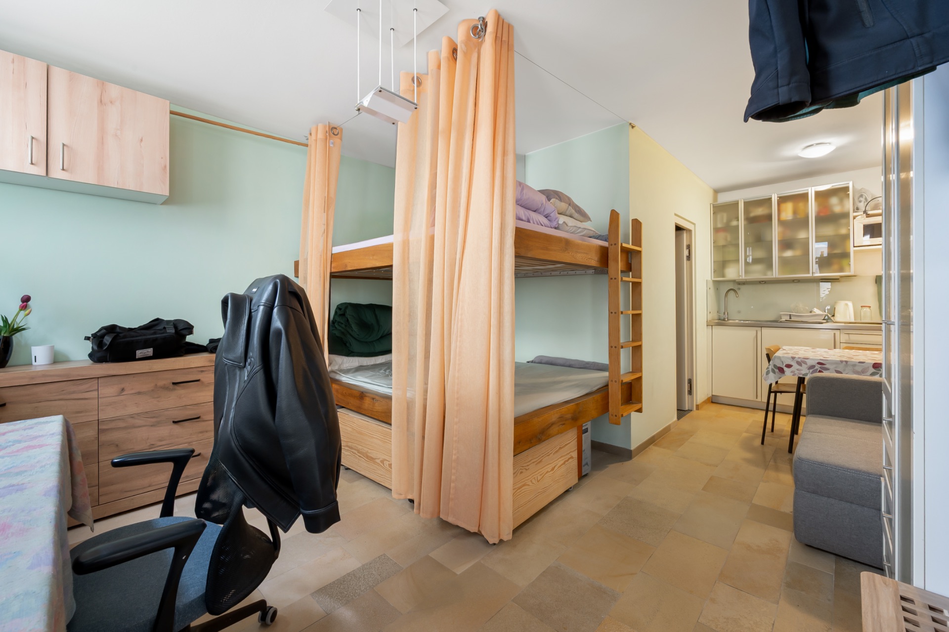 Zimmer mit Etagenbetten, Vorhängen, Schreibtisch, Stuhl und kleiner Küchenzeile im Hintergrund.