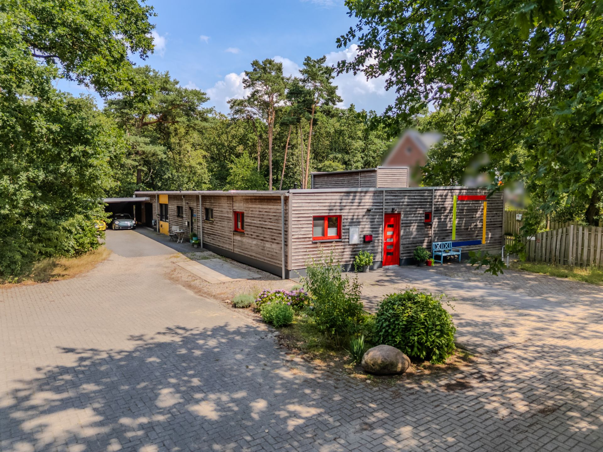 Modernes Flachbau-Holzhaus mit roten Fenstern, roter Tür und Vorgarten, umgeben von Bäumen.