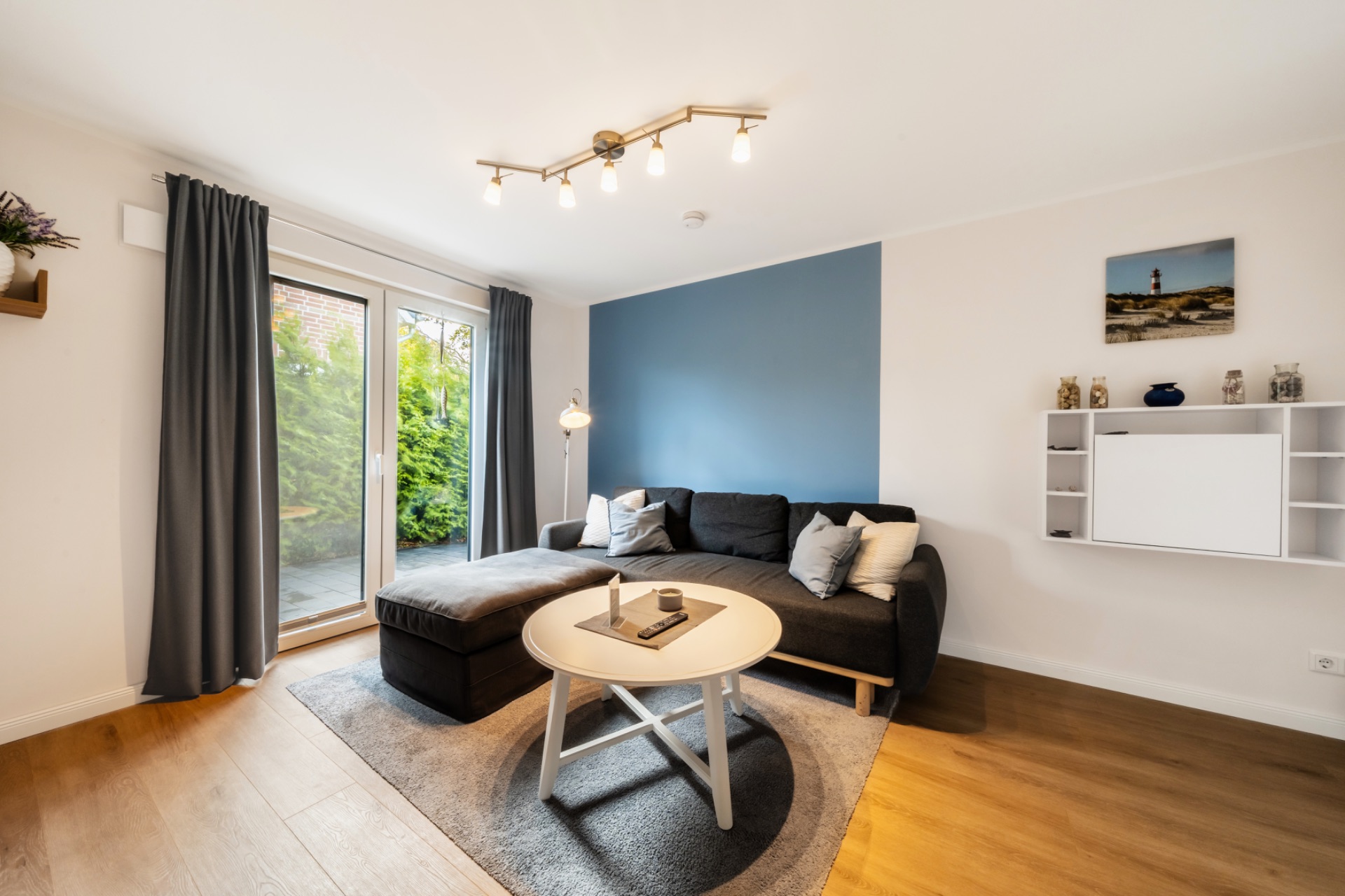 Helles Wohnzimmer mit Ecksofa, Couchtisch, Wandregal und großem Fenster mit Blick ins Grüne.