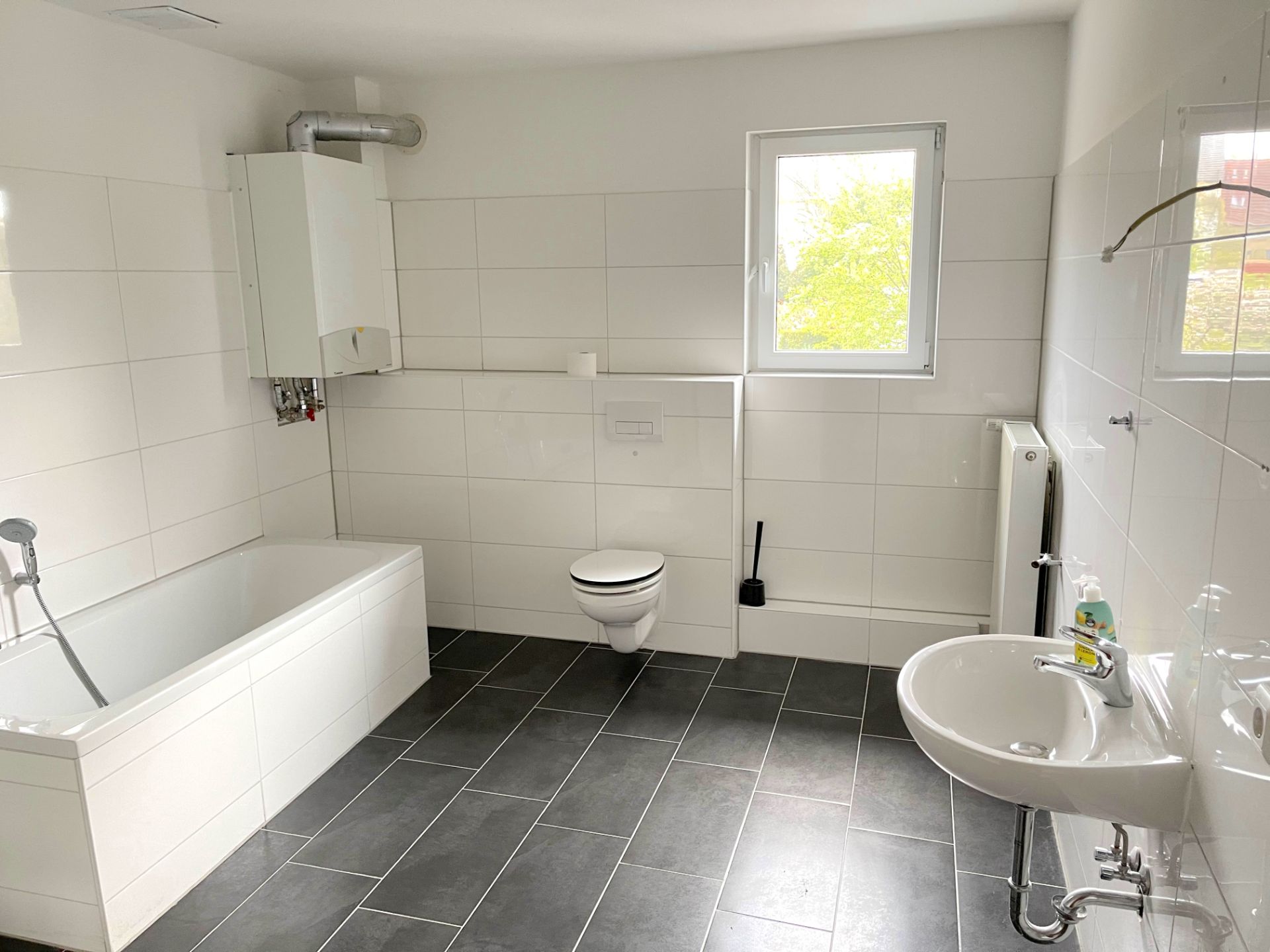 Helles Badezimmer mit Badewanne, Wandtoilette, Waschbecken und Fenster.