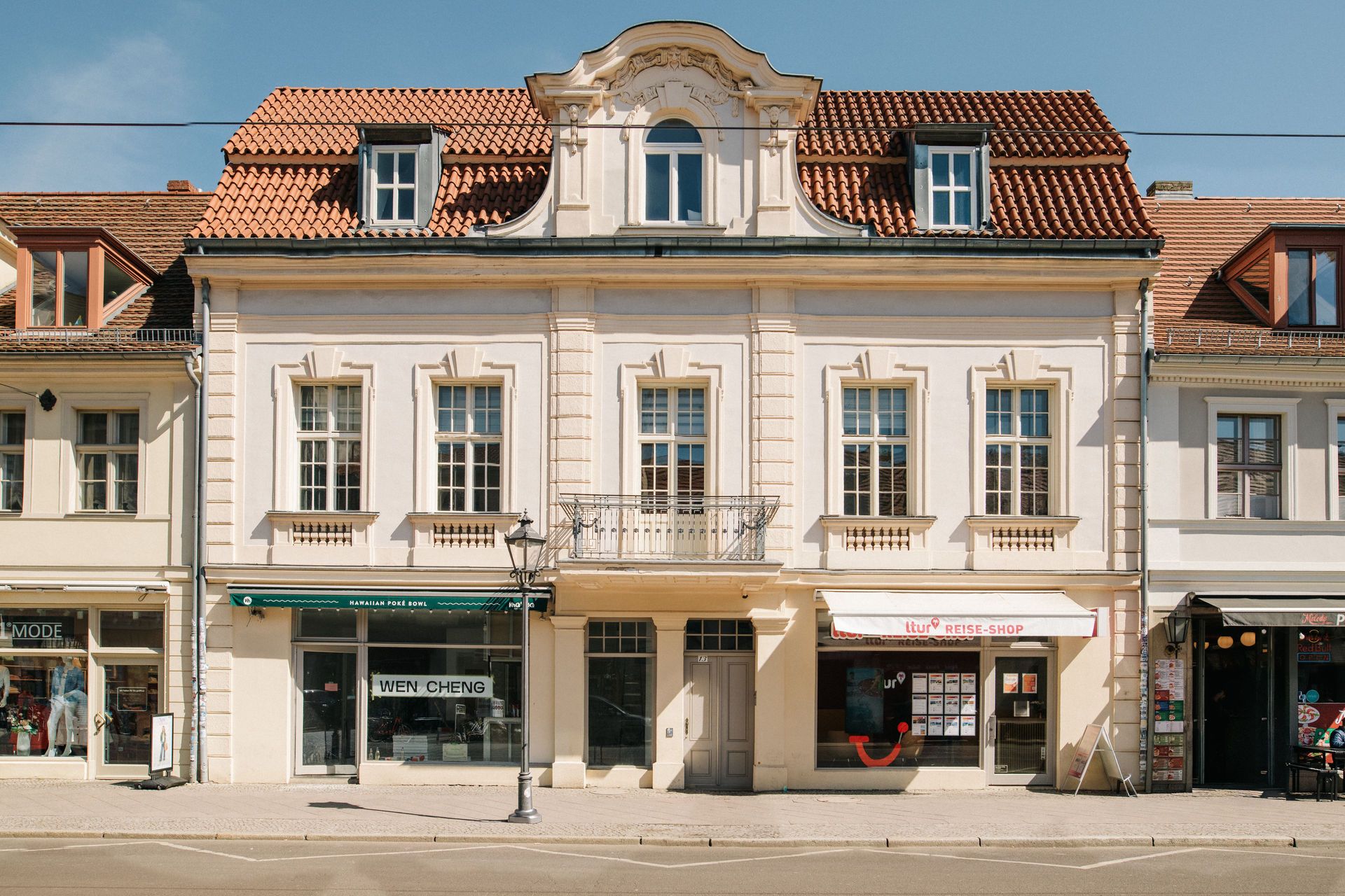 Fassade eines mehrstöckigen Wohnhauses mit Geschäften im Erdgeschoss und roten Dachziegeln.