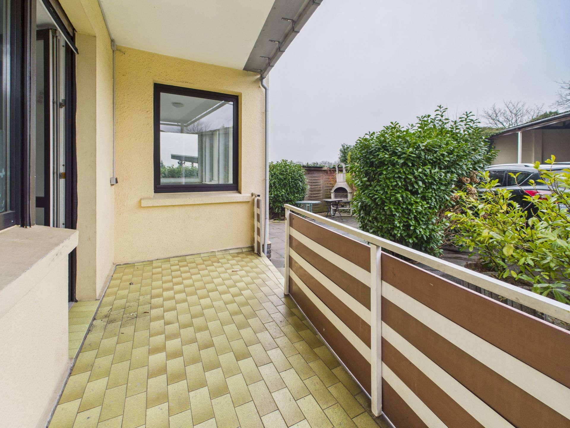 Überdachte Terrasse mit gelben Fliesen, Zaun und Blick auf Garten und Sträucher.