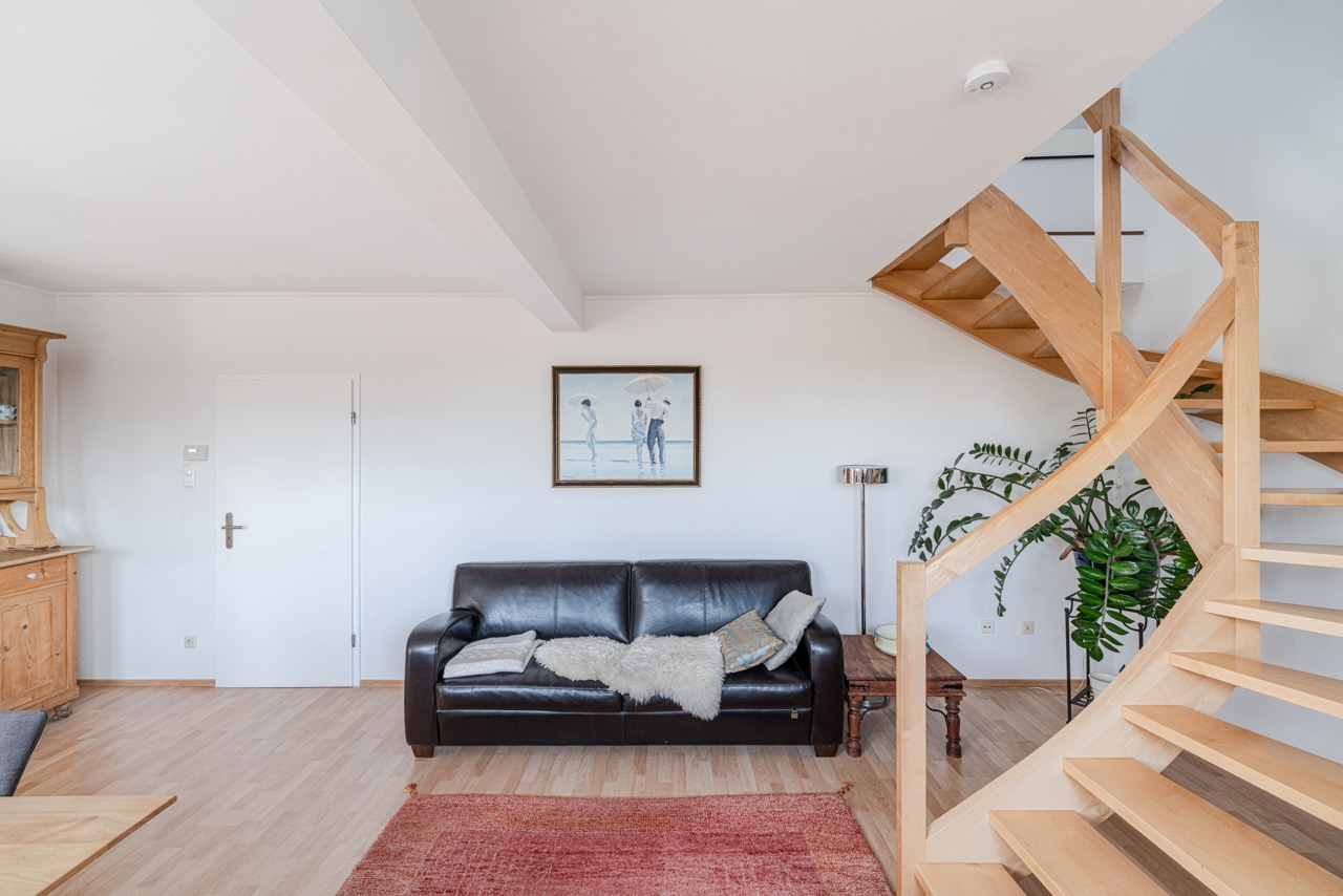 Wohnzimmer mit schwarzem Ledersofa, Holztreppe, Pflanze und Bild an der Wand.