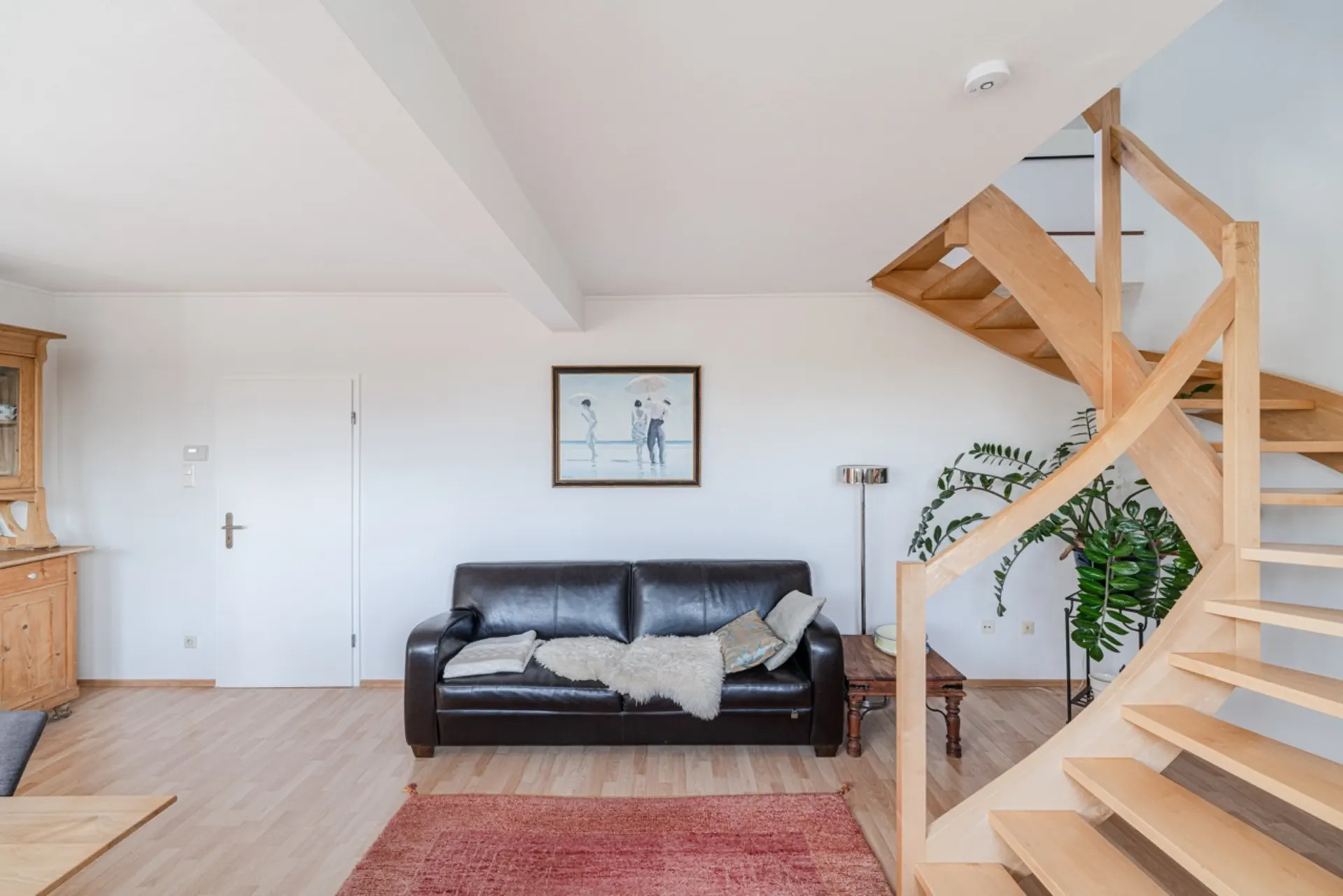Wohnzimmer mit schwarzem Ledersofa, Holztreppe, Pflanze und Bild an der Wand.