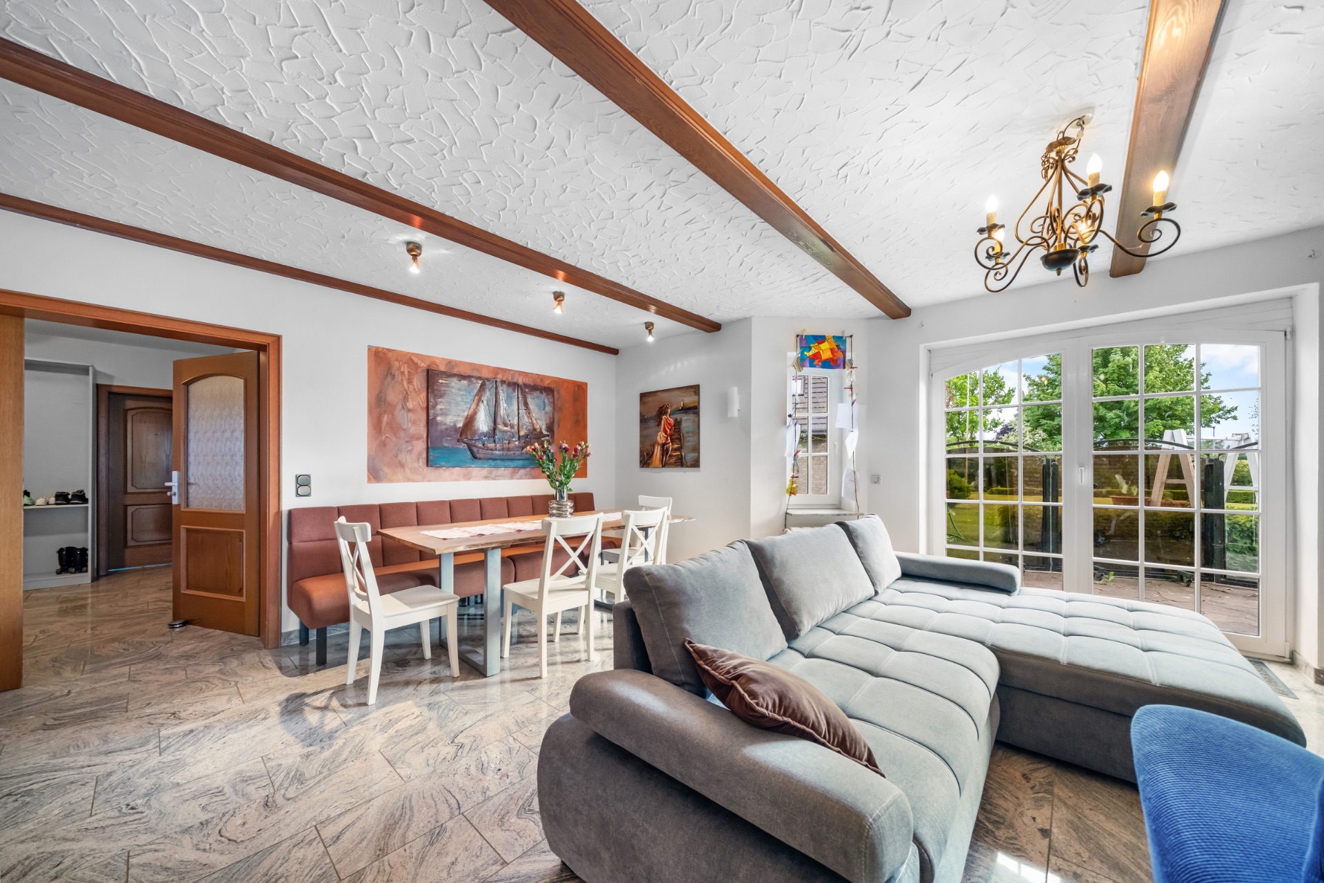 Helles Wohnzimmer mit Sofa, Esstisch, Stühlen, Gemälden und großen Fenstern mit Blick nach draußen.