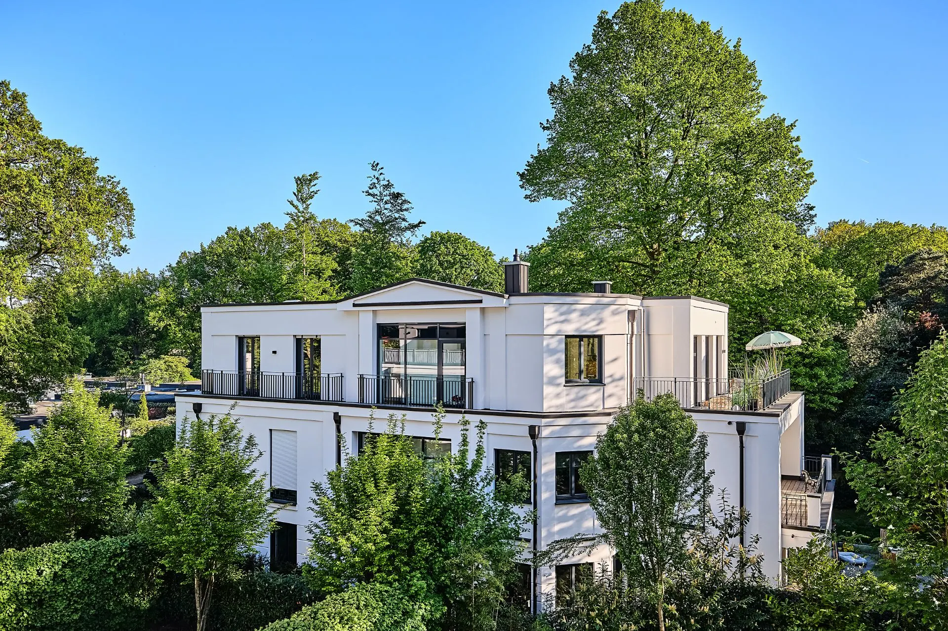 Modernes, weißes Wohngebäude mit Flachdach und großen Fenstern, umgeben von Bäumen.