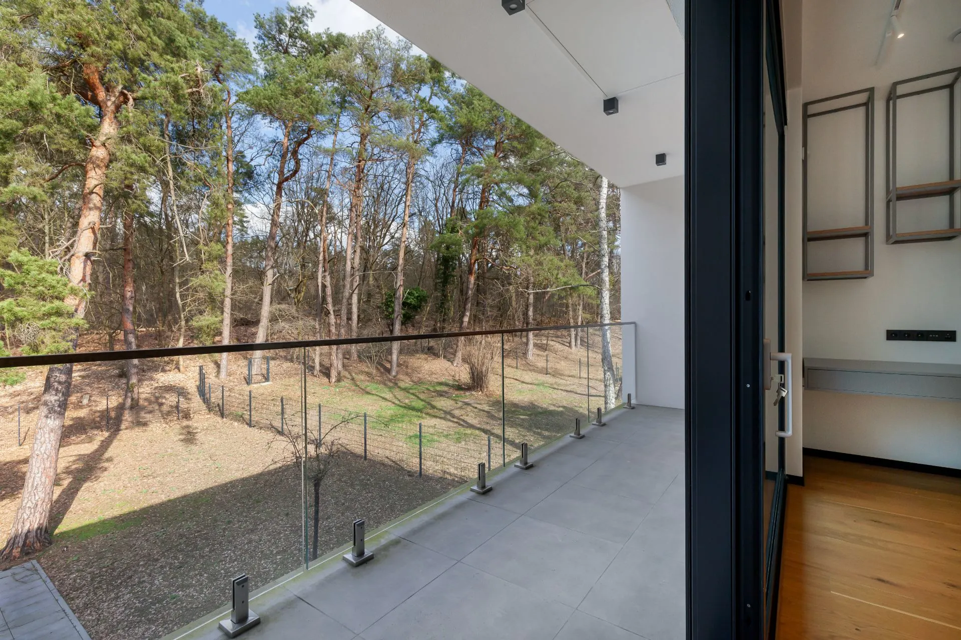 Großer Balkon mit Glasgeländer, Blick auf Bäume und Garten, angrenzend an einen Innenraum mit Holzboden.