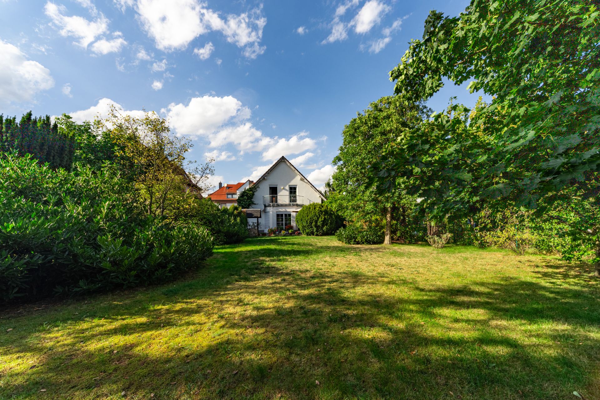 Weiße Wohnhausfassade mit großem Garten und vielen Bäumen an einem sonnigen Tag.