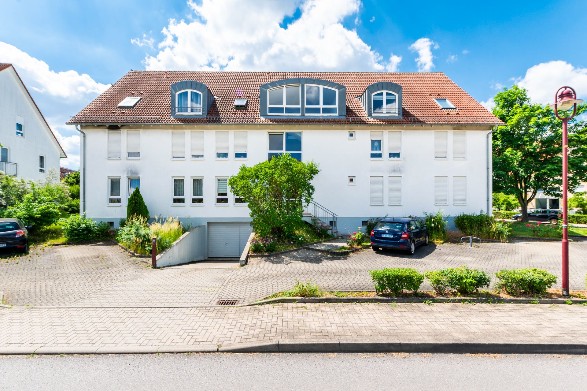Modernes Wohnhaus mit weißer Fassade, rotem Dach und Parkplätzen davor, einige Autos und Bäume sichtbar.