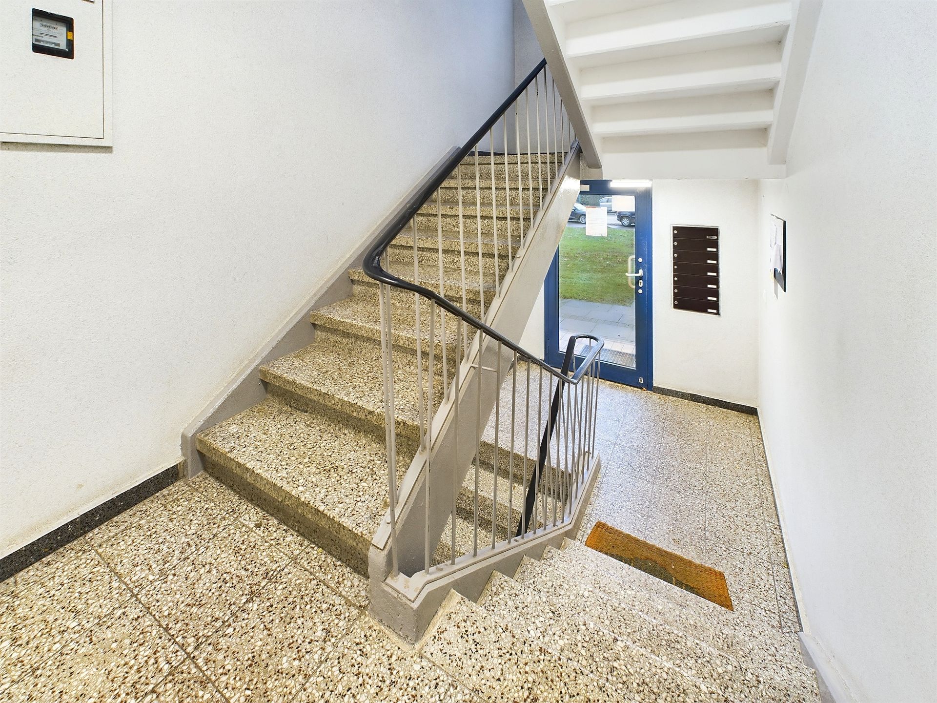 Helles Treppenhaus mit Geländer, Briefkästen und Ausgangstür in einem Wohngebäude.