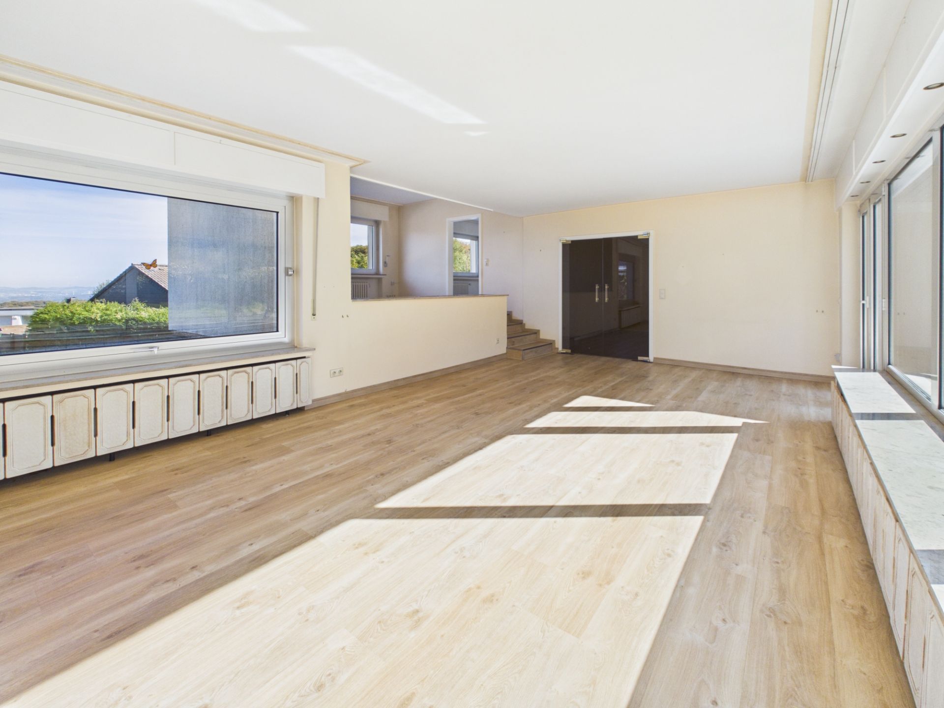 Helles, leeres Wohnzimmer mit großen Fenstern, Parkettboden und Blick nach draußen.