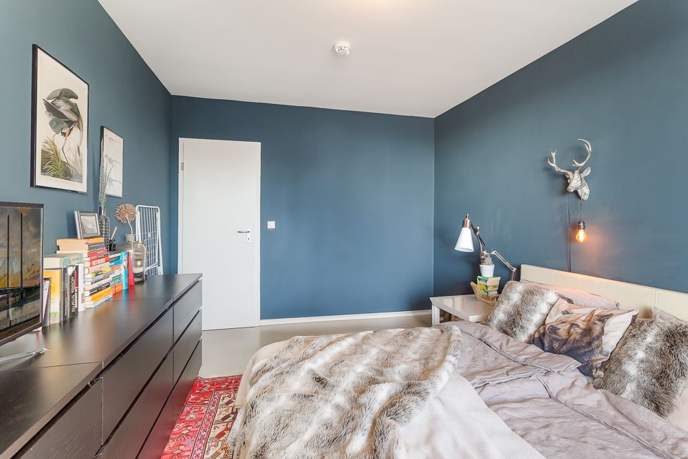 Modernes Schlafzimmer mit blau gestrichenen Wänden, Bett, Kommode mit Büchern und Wanddekoration.