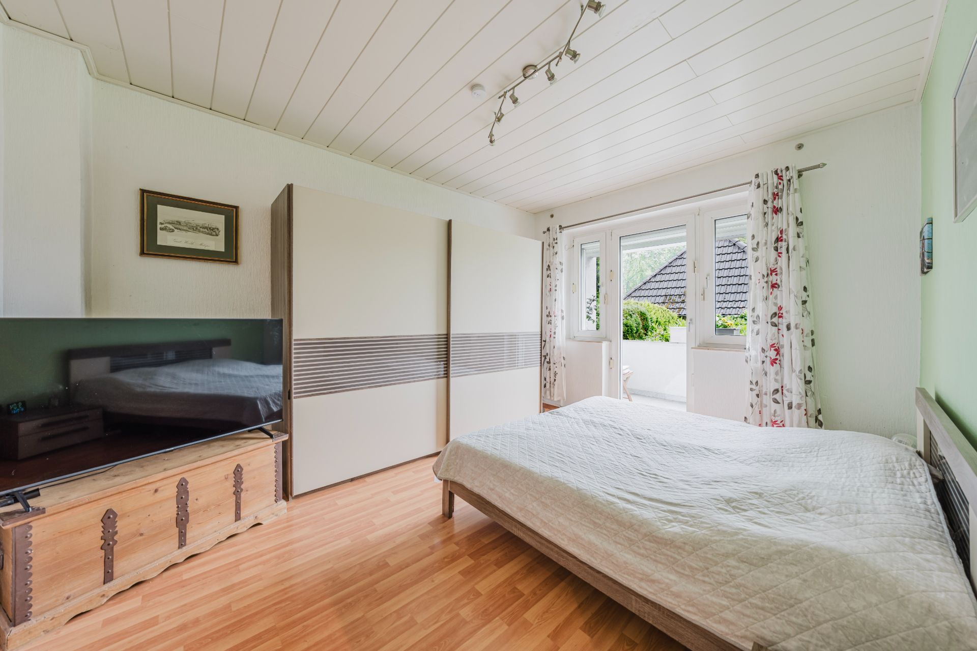 Schlafzimmer mit Doppelbett, Kleiderschrank, Fernseher und Zugang zu einem Balkon mit Vorhängen.