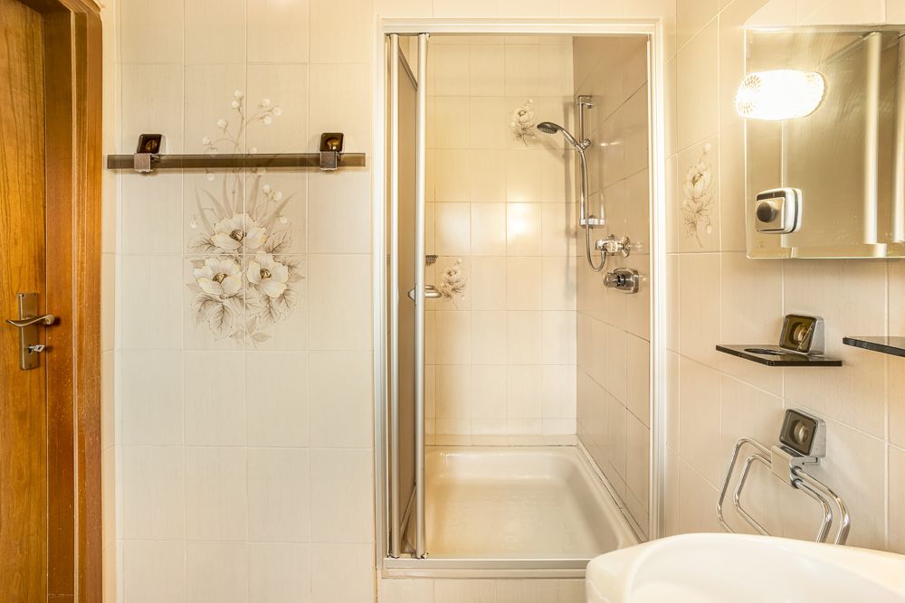 Helles Badezimmer mit Dusche, Spiegel, Waschbecken und Wanddeko mit Blumenmotiv.