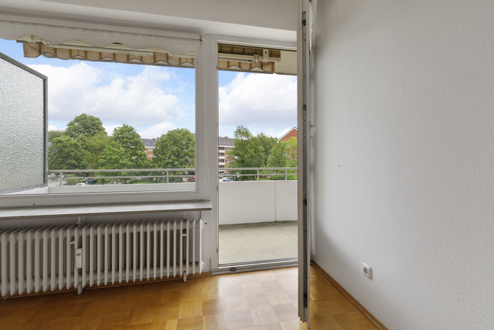 Heller Raum mit Parkettboden, Heizkörper und Zugang zu einem Balkon mit Blick auf Bäume.