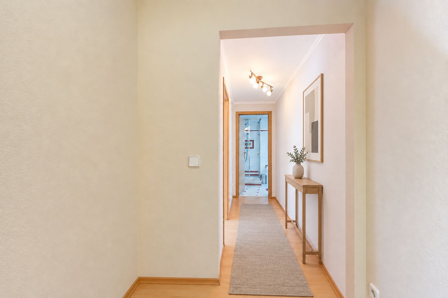 Heller Flur mit Teppichläufer, kleinem Tisch, Bild an der Wand und Blick ins Badezimmer.