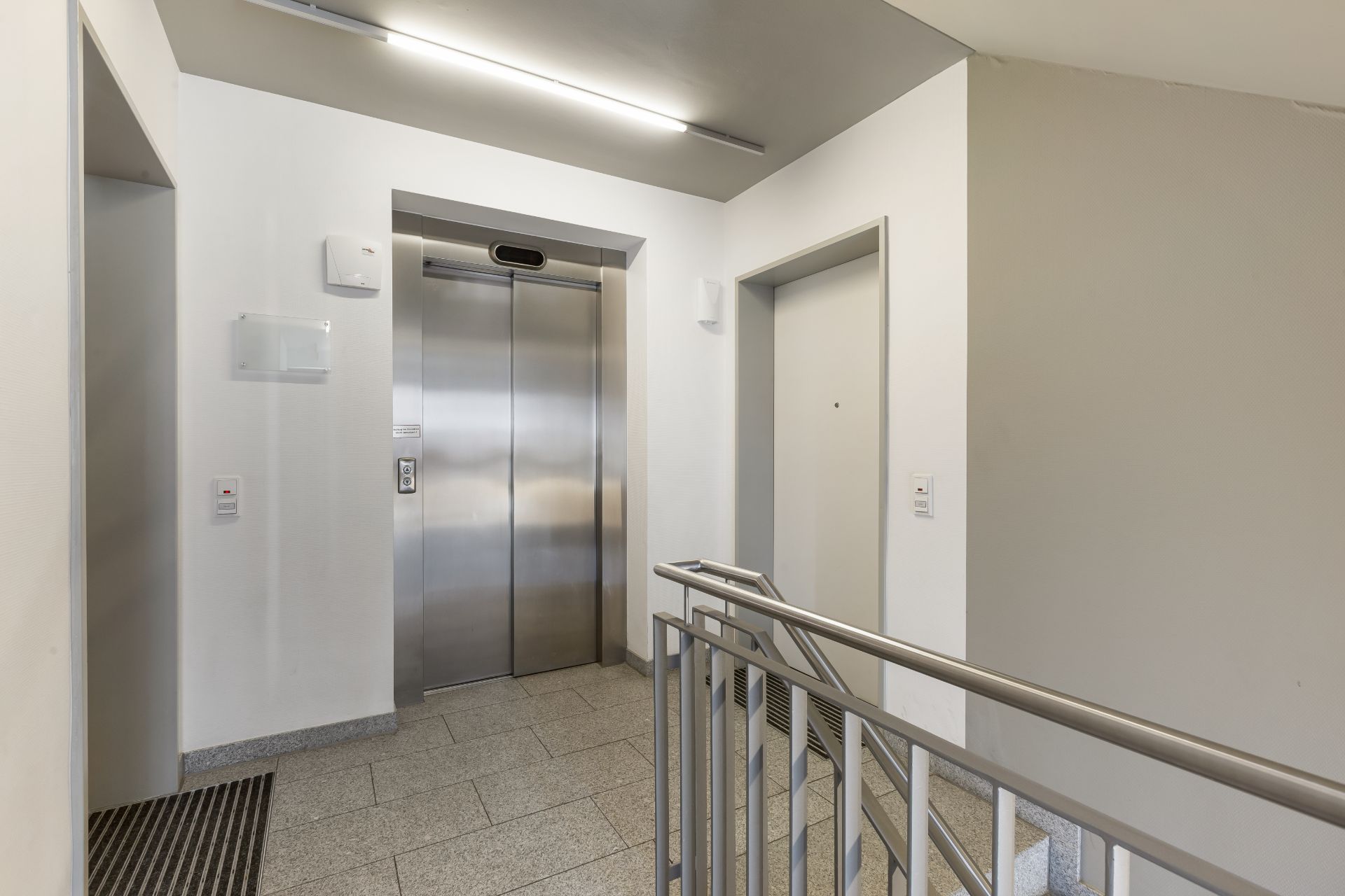 Heller Flur mit Aufzug, Treppengeländer und zwei Türen in einem Wohnhaus.