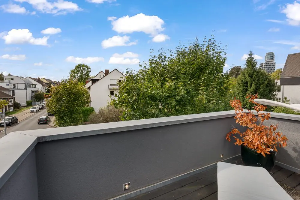 Balkon mit grauer Brüstung, Pflanze im Topf und Blick auf Bäume sowie Wohnhäuser.