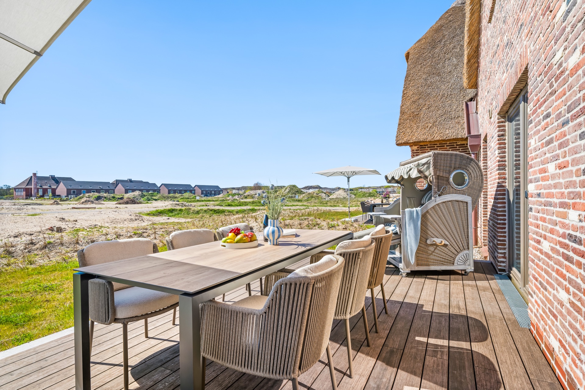 Terrasse mit Esstisch, Stühlen, Strandkorb und Blick auf andere Häuser im Hintergrund.