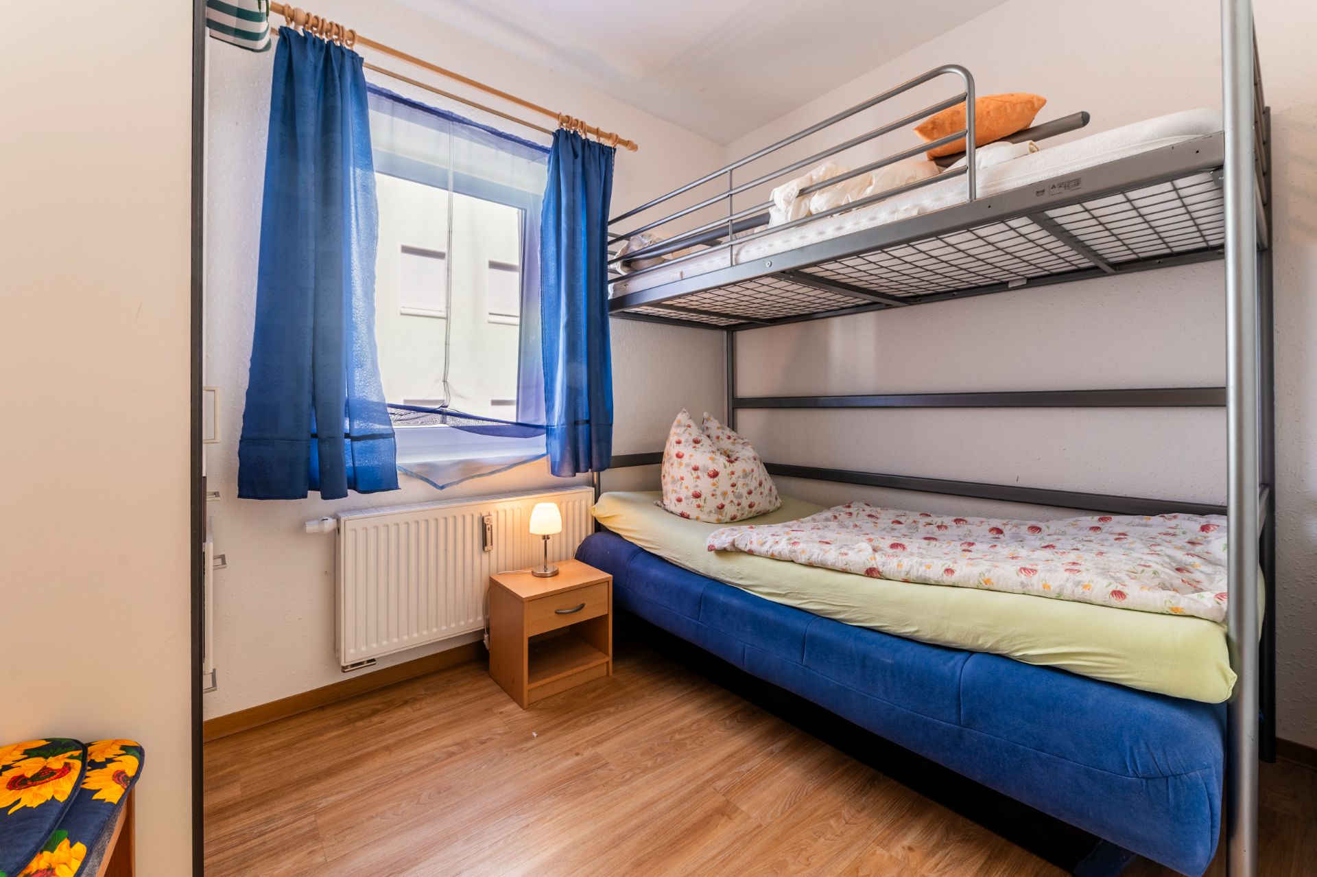 Kleines Schlafzimmer mit Etagenbett, Nachttisch und Fenster mit blauen Vorhängen.