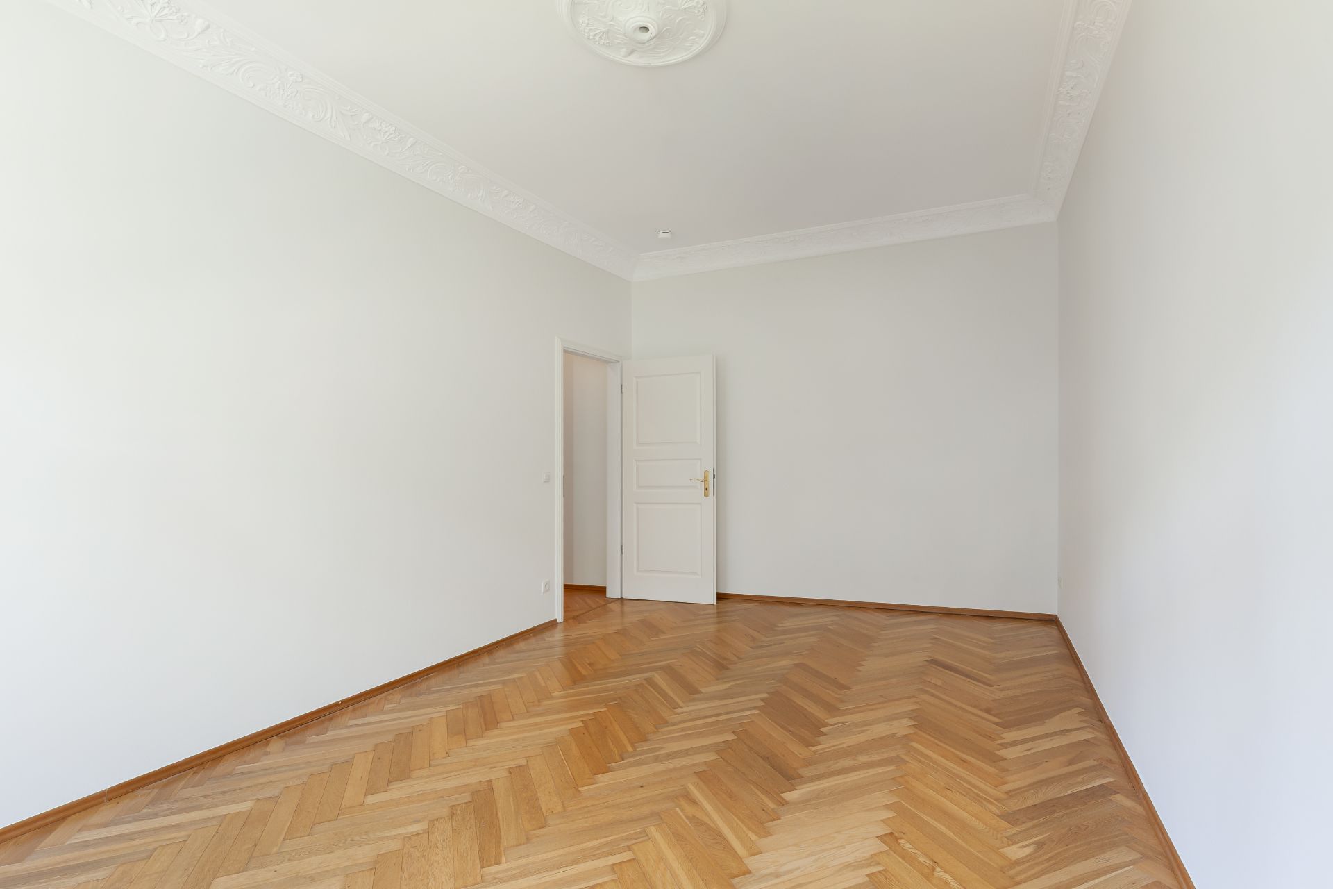 Leerer Raum mit Fischgrätparkett, weißen Wänden und geöffneter Tür.
