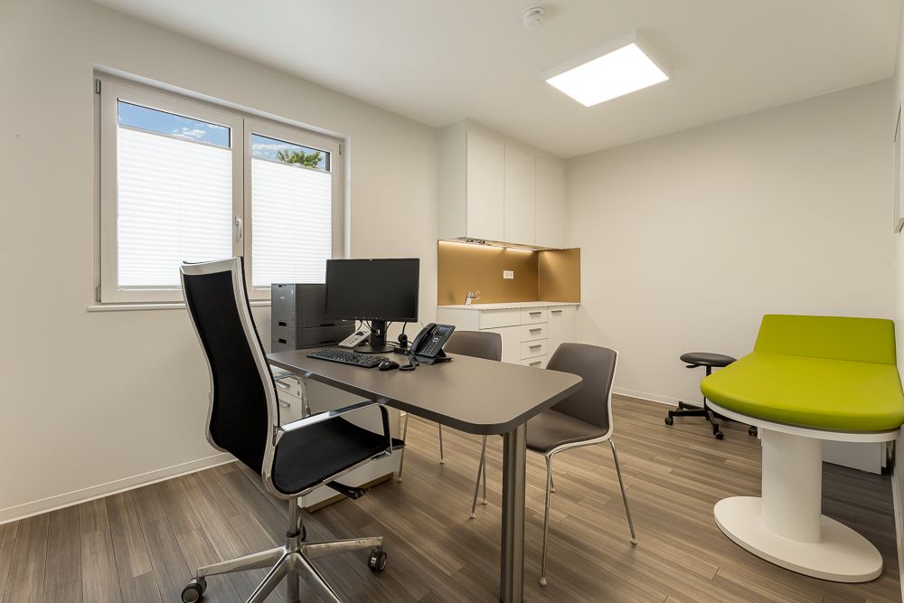 Modernes Büro mit Schreibtisch, Stühlen, Computer und grünem Sofa in hellem Raum.
