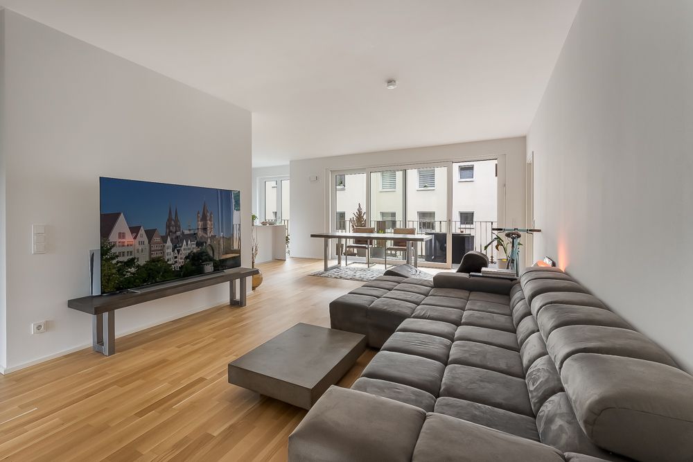 Modernes Wohnzimmer mit grauem Sofa, großem Fernseher und Zugang zum Balkon.