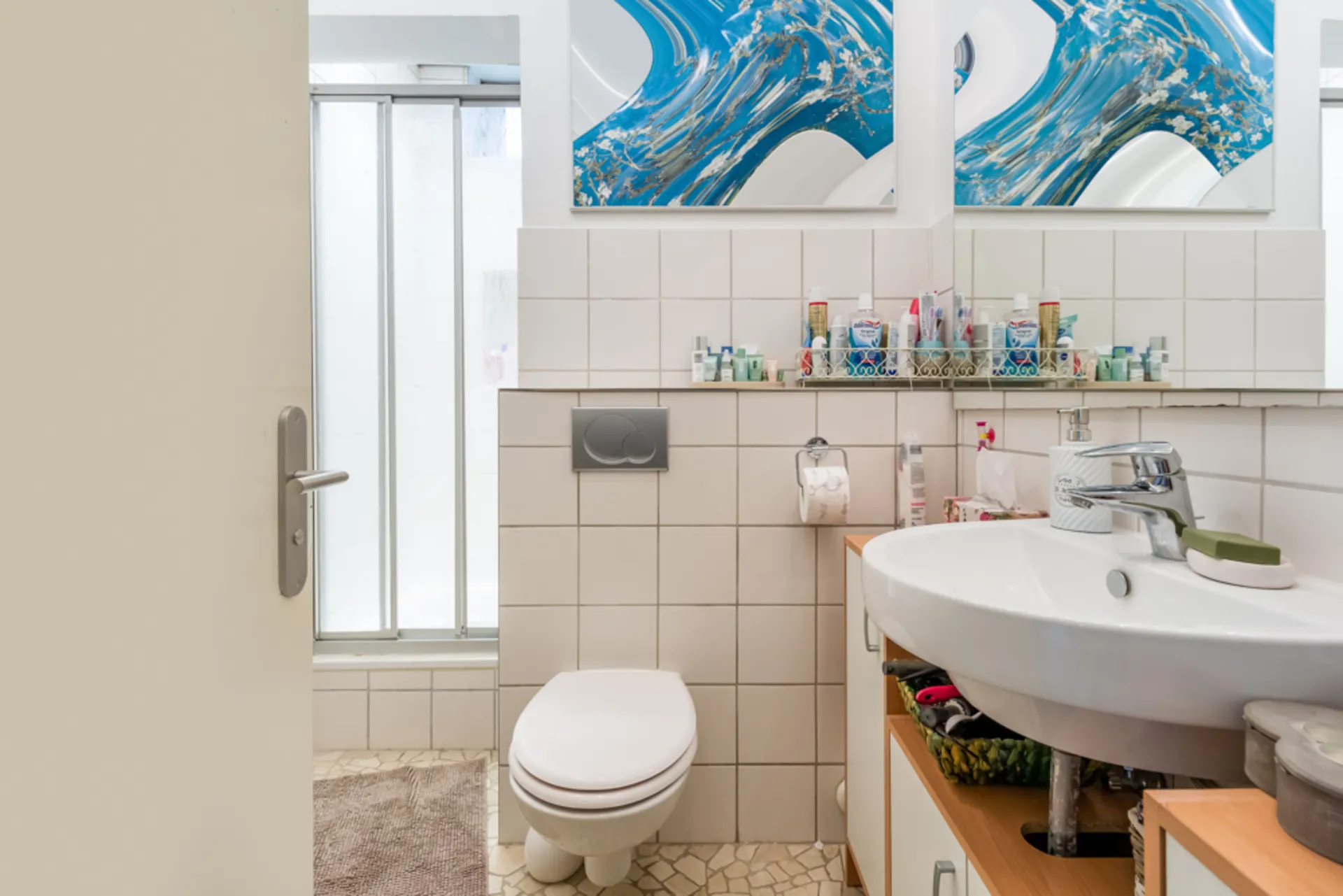 Helles Badezimmer mit Toilette, Waschbecken, Spiegel, Dusche und Pflegeprodukten auf einem Regal.