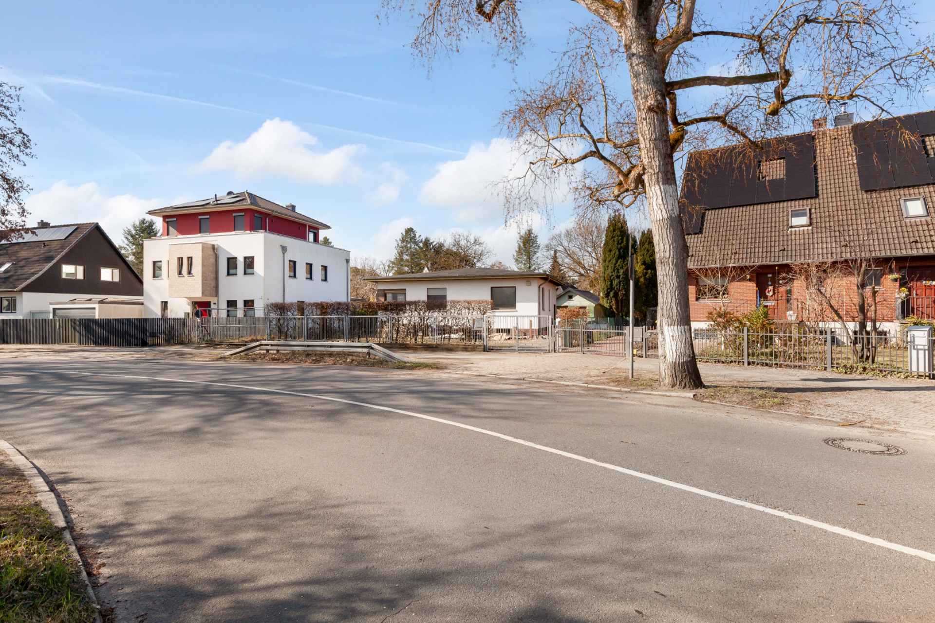 Straßenszene mit mehreren Wohnhäusern, Bäumen und einem Zaun im Vordergrund.
