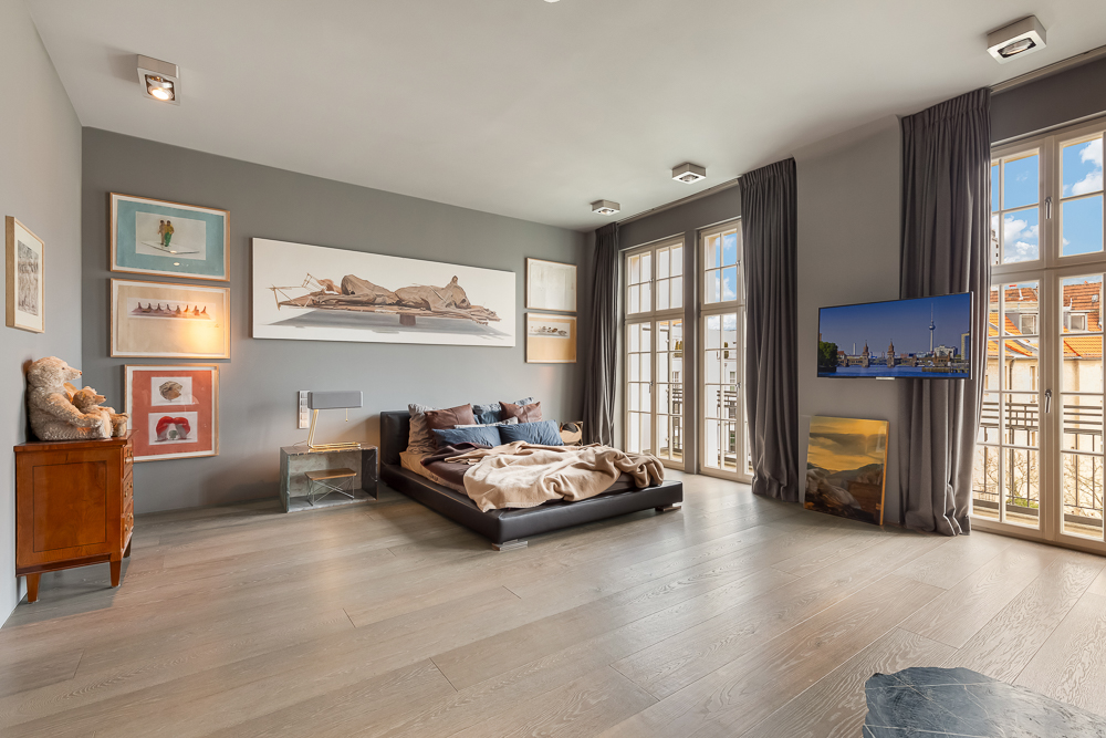 Modernes Schlafzimmer mit großem Bett, Holzboden, Bildern an der Wand und zwei großen Fenstern mit Gardinen.