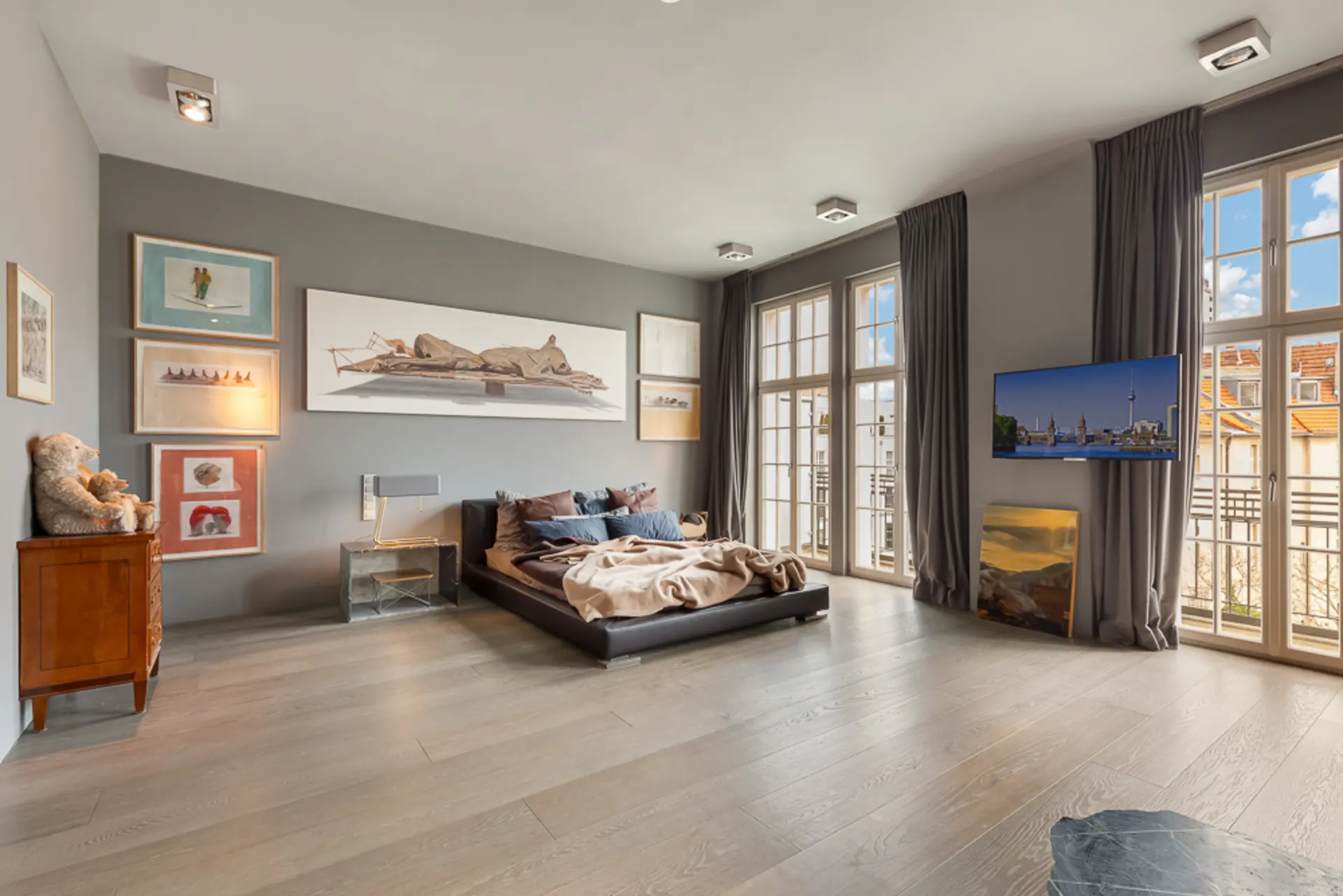 Modernes Schlafzimmer mit großem Bett, Holzboden, Bildern an der Wand und zwei großen Fenstern mit Gardinen.