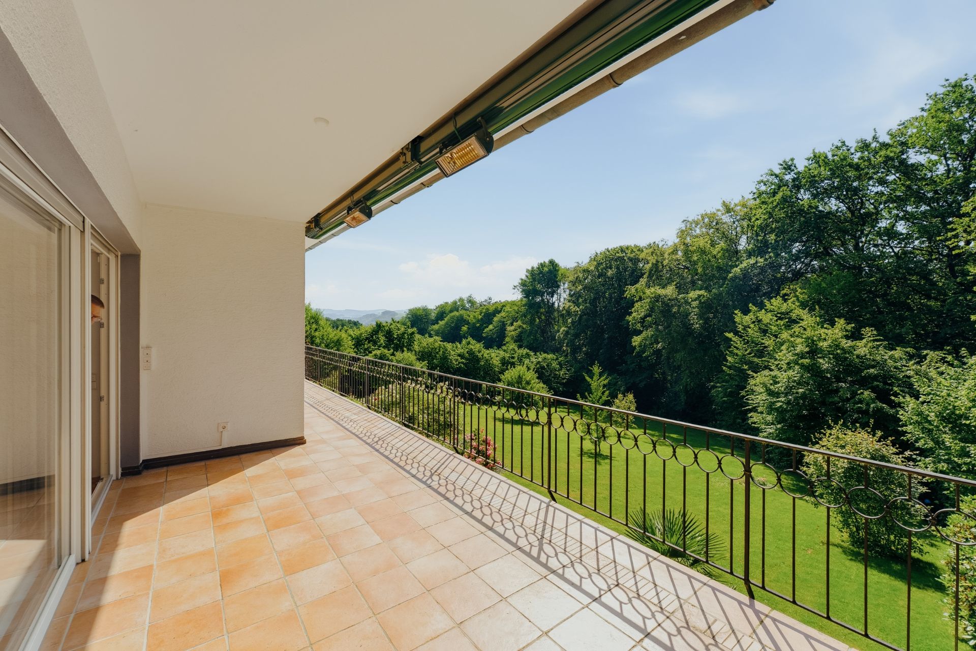 Große Terrasse mit Fliesenboden und Metallgeländer, Blick auf grünen Garten und Bäume.