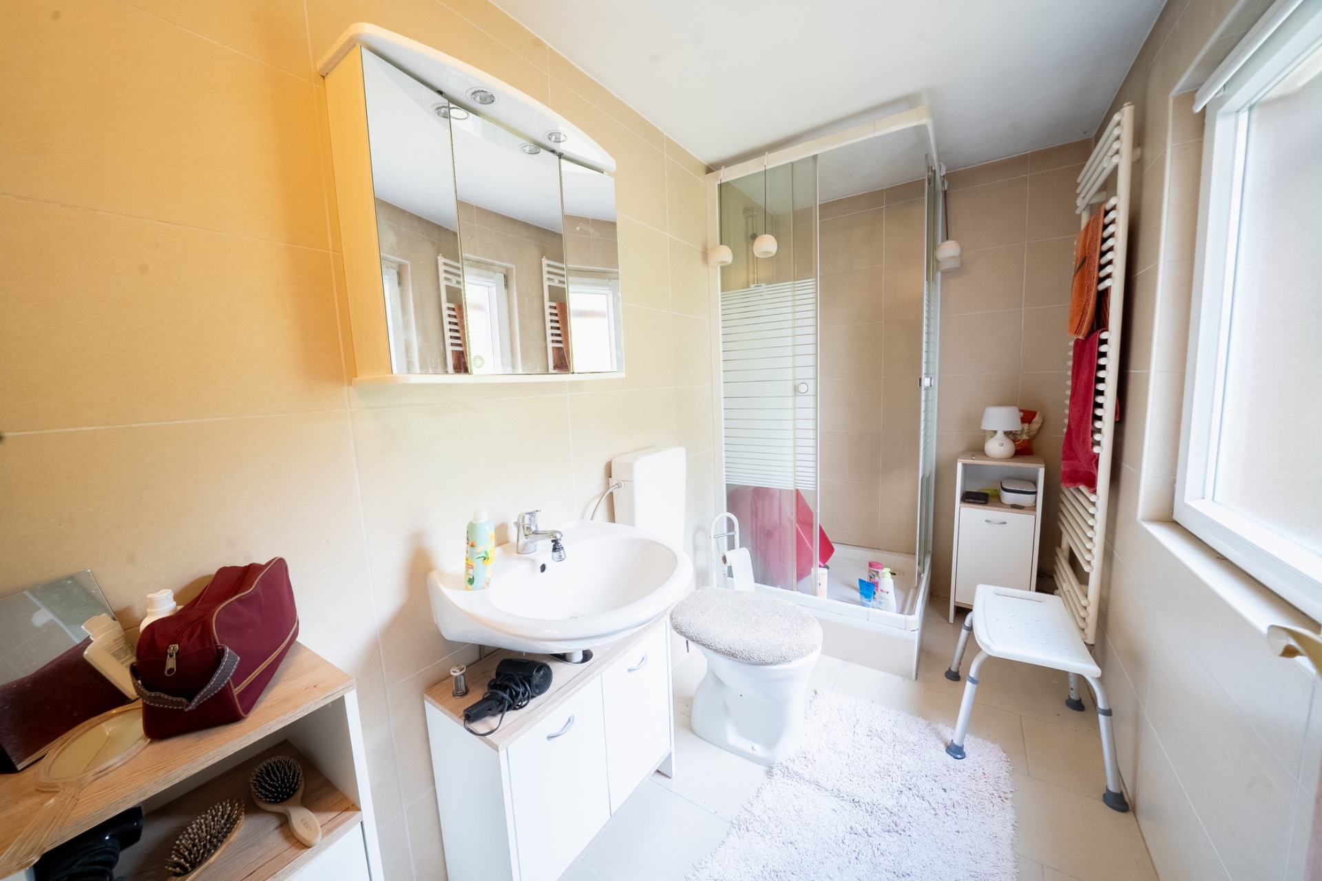 Helles Badezimmer mit Dusche, Waschbecken, WC und Regal, daneben ein Fenster und ein weißer Hocker.